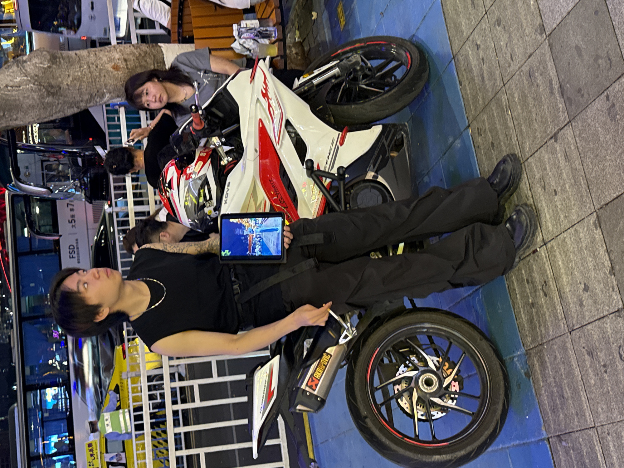 The scene shows an Asian man lying on a white and red motorcycle on the street. The man is wearing a black t-shirt, pants, and black shoes. He is looking up to his left. He holds a tablet computer. The exhaust of the motorcycle is branded with Akrapovic. Three other people, two women and one man, are laying near the motorcycle. The background includes city lights, vehicles, and a blue and gray street area. There is a yellow fence behind the man. The location appears to be in Yuzhong District, China.