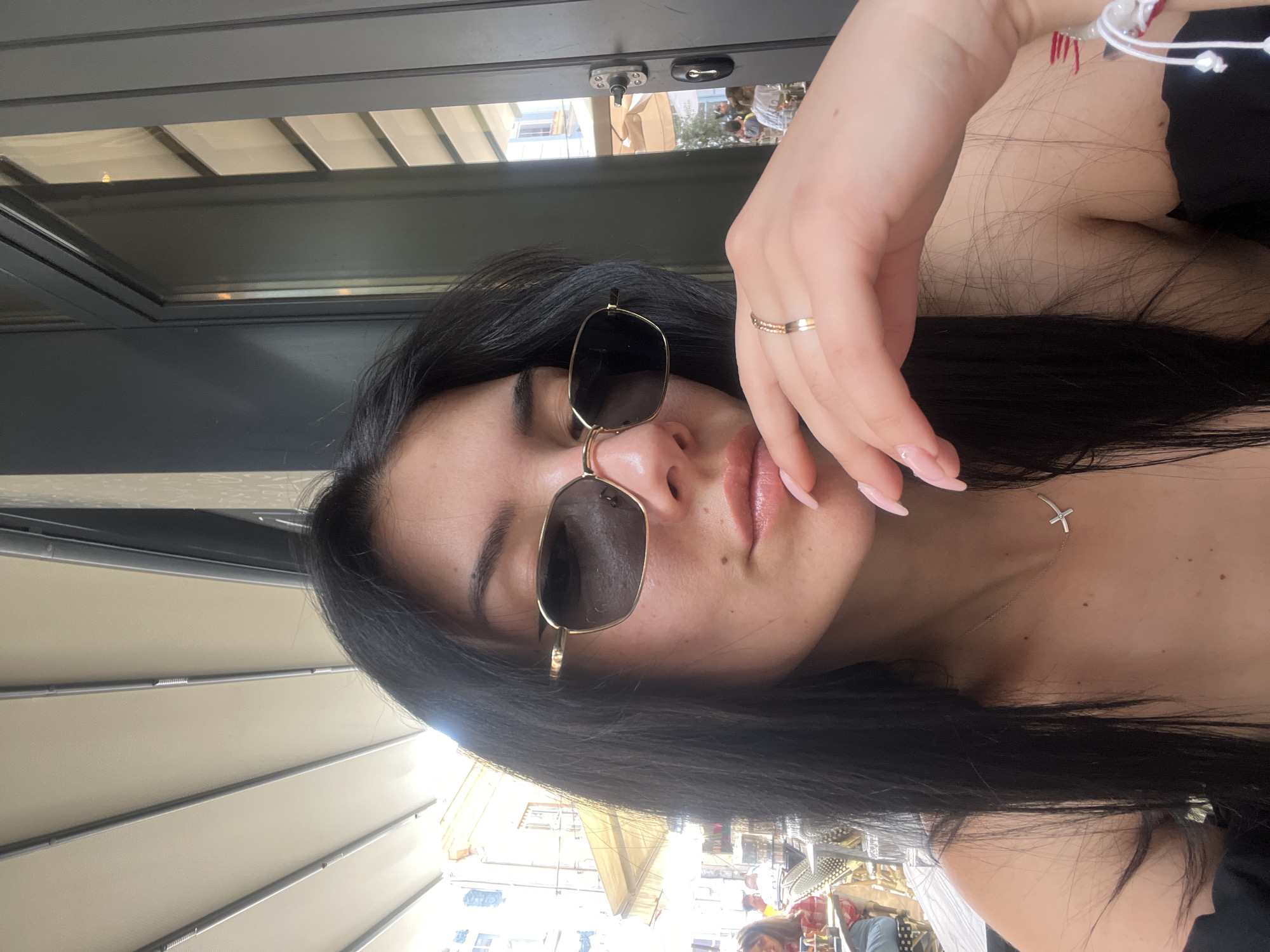 A young woman wearing sunglasses is pictured in a selfie taken outdoors in Plovdiv, Bulgaria. The shot appears to be at a restaurant or cafe. The woman has dark hair and a gold ring. A cross pendant is visible at her neck. The backdrop features sunshades and views of the city.