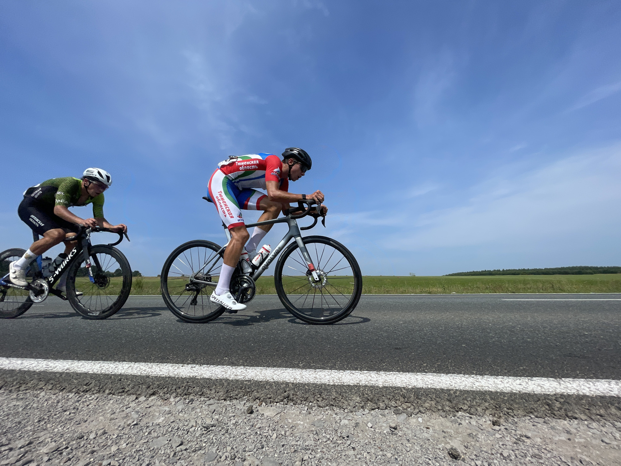 Two male cyclists are racing on a paved road under a bright blue sky with scattered white clouds. The cyclist in the foreground on the right wears a red, white, and blue racing suit with the inscription "Тюменская область" (Tyumen Oblast). He is riding a grey bicycle and is wearing a black helmet and white cycling shoes. The cyclist on the left wears a green and black racing suit and a white helmet. He is riding a black bicycle with "S-WORKS" visible on the frame and is also wearing white cycling shoes. The road has a white line marking and a gravel shoulder. In the background, there is a flat, green landscape with a treeline in the distance. The scene appears to be taking place in or around the village of Perevoznoe, Tyumen Oblast, Russia, based on the inscription on the cyclist's suit.