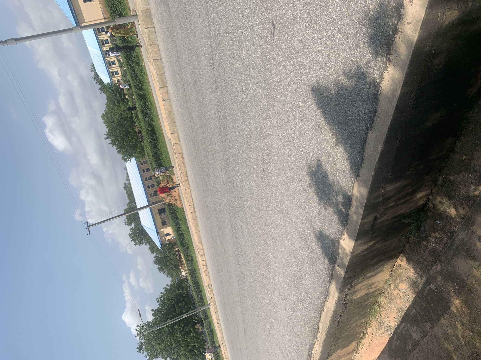 In Jalingo, Nigeria, an eye-level shot captures a wide asphalt road with a slight tilt to the left, suggesting it is a moderately trafficked area. The road surface is well-maintained, with visible tire marks and a clear demarcation line on the right side. A concrete curb runs along the left edge of the road, separating it from a sandy embankment. Overcast skies with scattered clouds loom above, casting dappled shadows of trees onto the road.
On the far side of the road, a row of buildings with light-colored walls and distinctive blue roofs suggests an institutional or educational facility. A manicured hedge runs along the front of the buildings, creating a neat boundary. Several people are visible in the distance, walking along the pavement in front of the buildings. One individual in a red shirt walks away from the camera, while others in traditional attire stroll along the walkway. A utility pole with power lines is positioned to the right of the frame, reinforcing the urban setting. The overall impression is one of a serene, functional environment, likely a university campus or government compound.