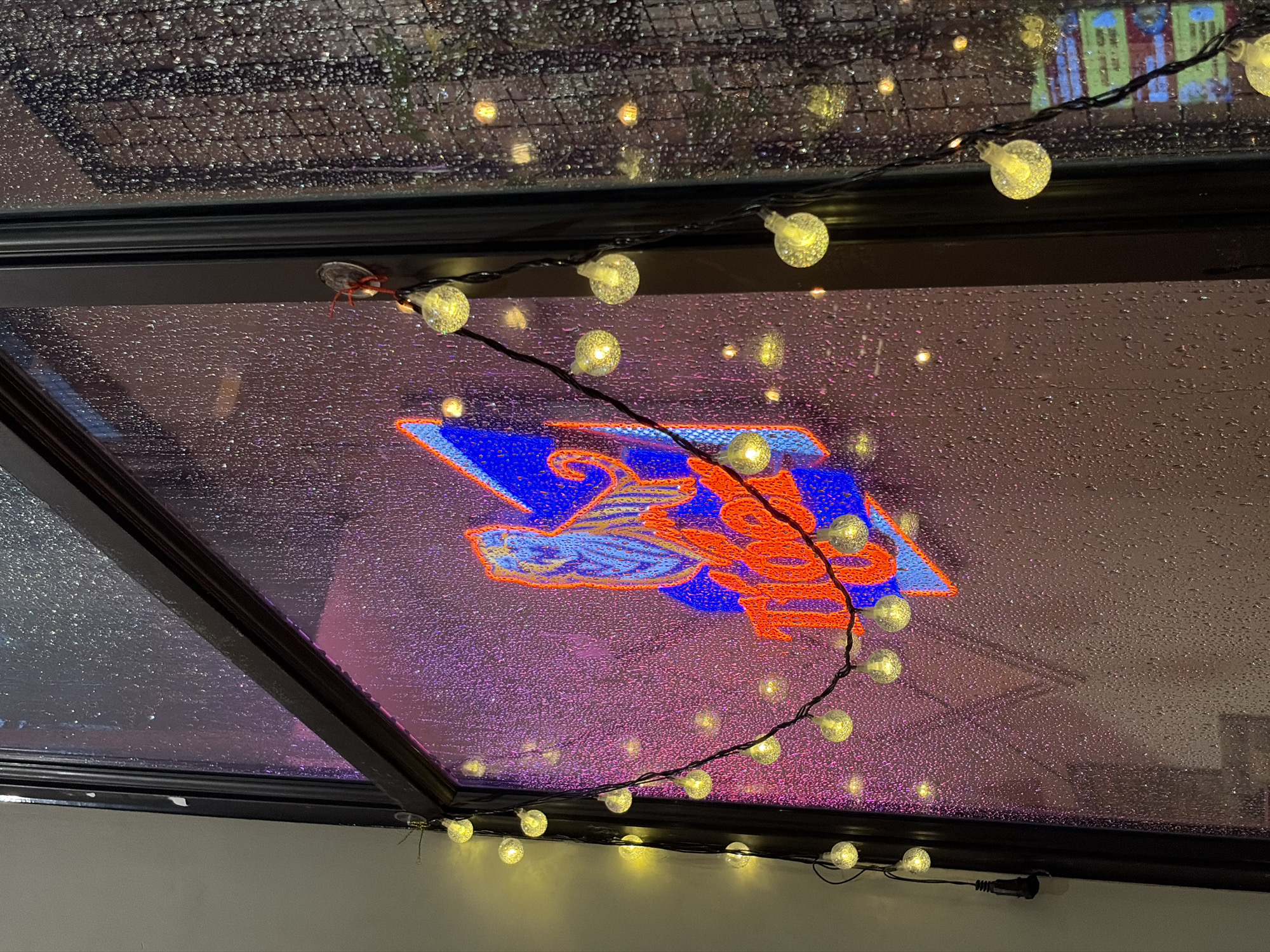 This image depicts a neon sign for "Tiger" beer, featuring a stylized tiger's head and the brand name in red and blue, displayed on a window. The window is covered in raindrops, creating a blurred, textured effect. A string of small, clear, globe-shaped lights is draped across the window, with some lights reflecting on the wet surface. The background appears to be a dark interior with some indistinct lighting and objects. The scene is likely in Thủ Đức, Vietnam, given the presence of Tiger beer, a popular brand in the region. The lighting and the rain suggest it is evening or nighttime. There are no people visible in the image.