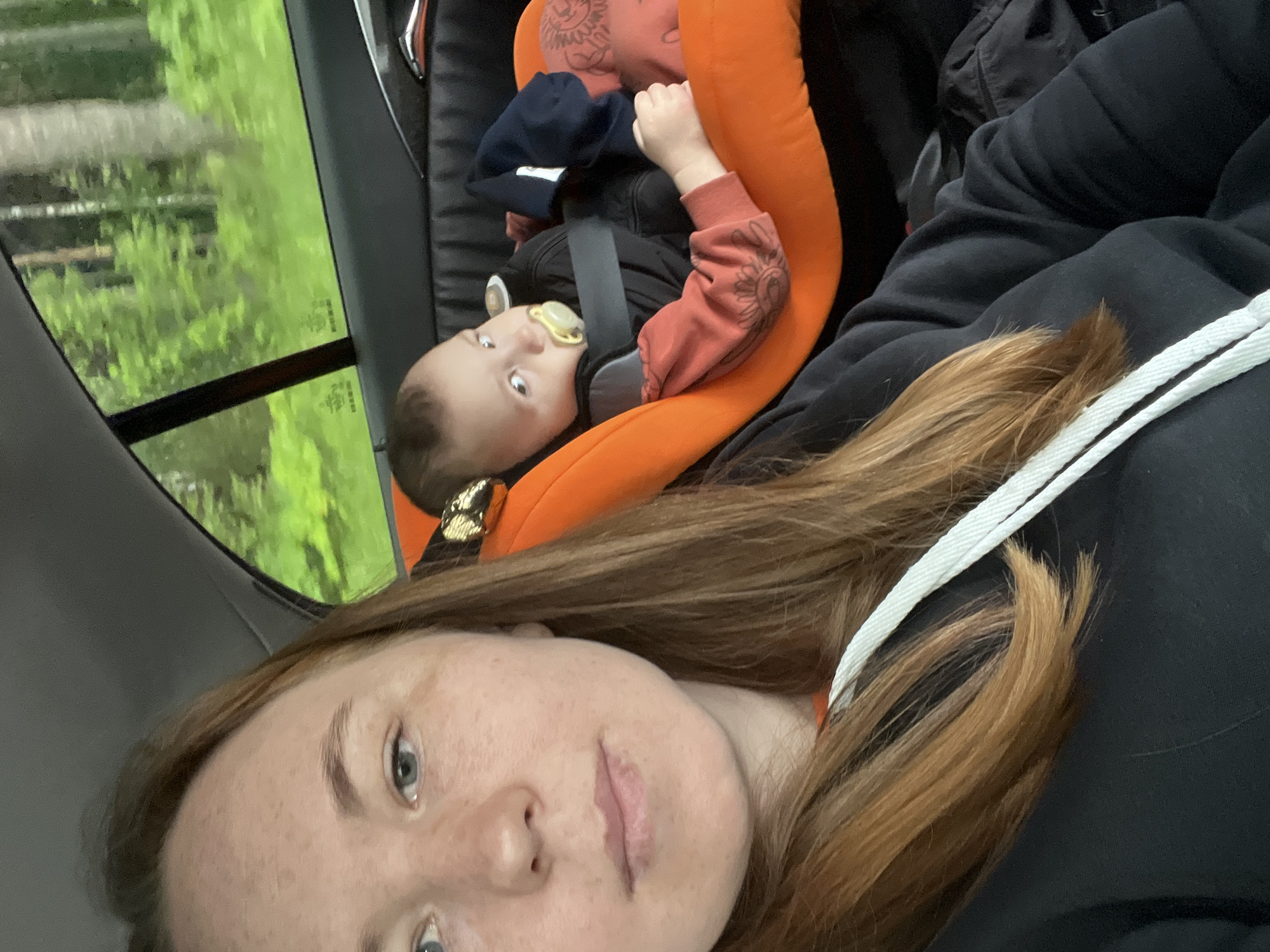 An adult female and an infant are depicted inside a vehicle. The female, positioned in the left foreground, has reddish-brown hair, light eyes, and freckles, and is wearing a black hoodie with white drawstrings. She looks directly towards the camera. In the right background, an infant or young toddler is secured in an orange car seat with grey inserts, wearing a dark long-sleeved top and reddish-orange patterned pants, with a yellow pacifier in its mouth. A dark blue fabric item is also present in the car seat. The car interior includes a grey headliner, dark paneling, and a metallic door handle partially visible on the right. Through the car window, a blurred green landscape of dense trees and foliage indicates a natural, possibly forested, environment. The scene takes place during daylight hours in the city of Прикамский, Russia.