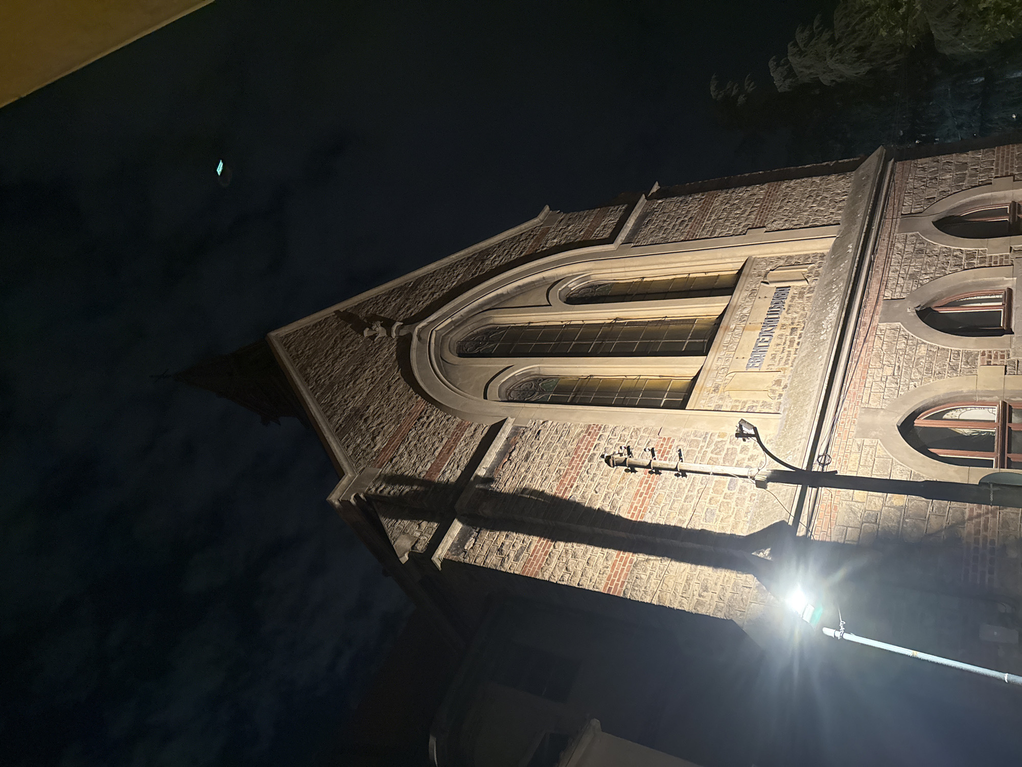 The image depicts a building at night in Plovdiv, Bulgaria. The building has arched windows and is constructed of brick and stone. A street light is visible in the foreground, casting a shadow on the building. The sky is dark and cloudy. A sign can be identified on the building.