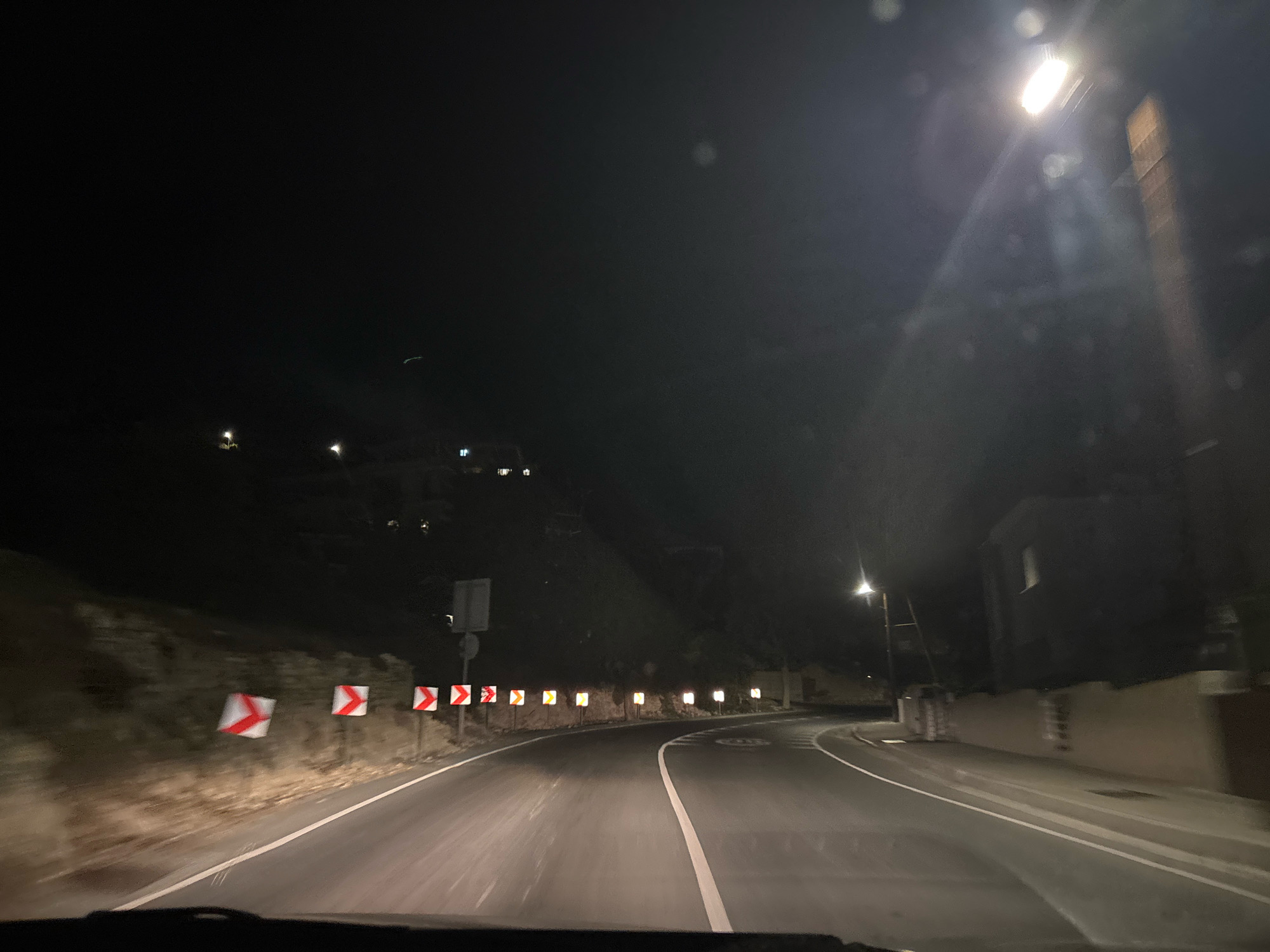 The image depicts a nighttime view of a winding road in Agios Tychona Community, Cyprus. The road is paved and has white lane markings. Numerous reflective warning signs, predominantly red and white chevrons indicating curves in the road, are placed along the roadside. A street light illuminates the scene from the right side of the frame. In the background, a hillside is visible with some scattered lights, suggesting buildings or settlements. The overall environment appears to be rural or semi-rural, with dense vegetation on the hillside. The image is captured from the perspective of a vehicle, as indicated by the dashboard visible at the bottom of the frame. The sky is dark, indicating it is nighttime.