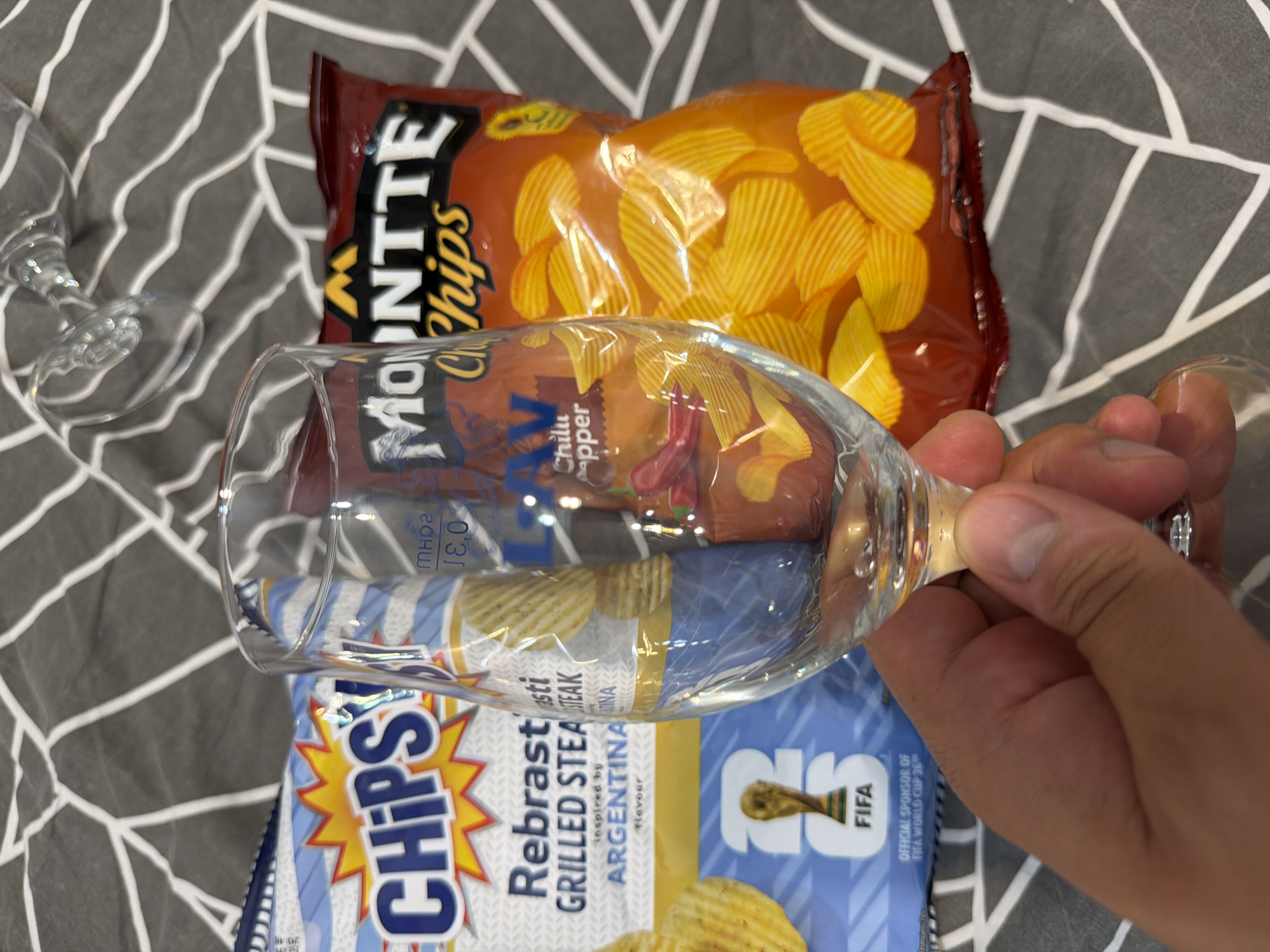 A person's hand holds a clear wine glass filled with red chili peppers and what appears to be a small bottle or packet. The glass is positioned in front of two bags of potato chips on a grey and white patterned surface. The bag on the left is blue and white with the brand "CHIPSY" prominently displayed. Text on this bag includes "Rebrast" and "GRILLED STEAK" with "ARGENTINA" and the FIFA World Cup trophy logo. The bag on the right is red and features the brand "MOUNTE Chips" with "Chili Pepper" text. The background suggests an indoor setting.