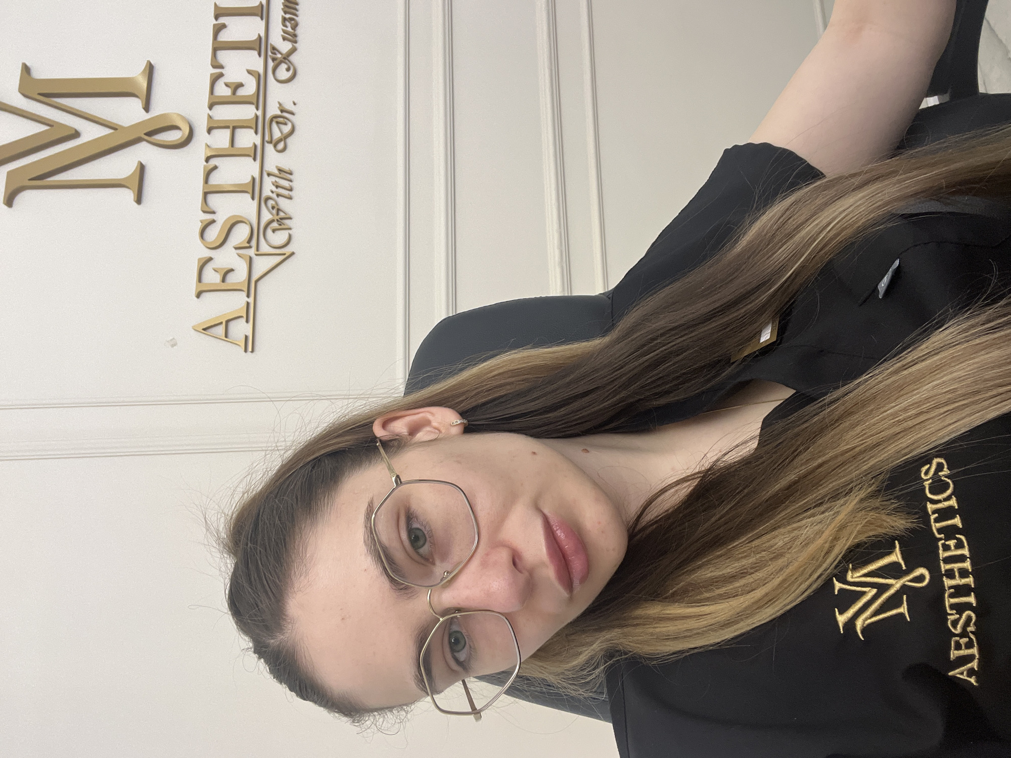 The image shows a close-up, vertically oriented selfie of a young woman. She has long, brown and blonde highlighted hair that falls over her shoulders and chest. She is wearing glasses with hexagonal frames and appears to be looking towards the camera with a neutral expression. Her lips are slightly parted and have a subtle gloss. She is wearing a black top with gold embroidery. The embroidery on her top features a stylized "M&N" logo and the word "AESTHETICS" in gold lettering.
Behind her, a white wall with vertical paneling is visible. To the left of the woman, a gold metallic 3D sign is mounted on the wall. The sign displays "M&N" in large, ornate lettering, followed by "AESTHETICS" and then "With Dr. Kuzma".
The lighting appears to be soft and even, suggesting an indoor setting. There are no clear indications of the time of day or weather conditions. The overall scene suggests a professional or clinical environment, likely related to aesthetics or medical practice, given the signage and her attire. The woman's appearance is neat and professional.