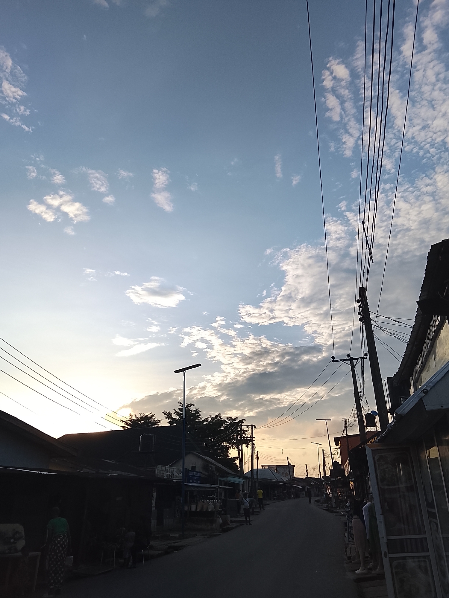 The image depicts a street scene in Rumuepirikom, Nigeria, during what appears to be late afternoon or early evening. The sky is a pale blue, partially filled with scattered white and grey clouds, with the sun's glow visible near the horizon on the left side of the frame. Overhead, multiple black electrical wires stretch across the sky, supported by wooden utility poles.
The street itself is paved and curves slightly into the distance, lined on both sides by buildings and small shops. The buildings exhibit a range of architectural styles, with some appearing to be single-story structures with corrugated metal roofs, while others are taller with more traditional construction. Many of the shops have open fronts, displaying goods for sale, including clothing on mannequins on the right side of the street.
Several individuals are visible in the scene. On the left, a person in a green top and patterned skirt walks away from the camera. Further down the street, other figures are seen walking or standing outside the shops. The overall atmosphere suggests a bustling commercial area. Trees are sparsely interspersed along the street, some with dark foliage. The lighting indicates a time of day when shadows are lengthening.