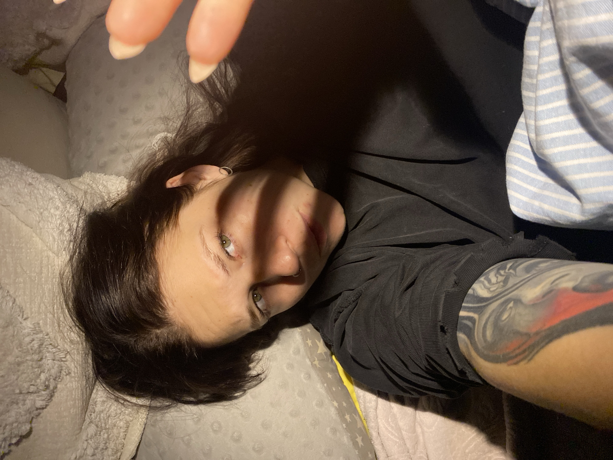 An overhead, slightly rotated close-up captures a person, likely female, lying down indoors in what appears to be Chaikovsky, Russia. The individual, with dark hair and striking green eyes, gazes directly towards the camera. Strong, linear shadows fall across the face, originating from what appears to be fingers with long, light-colored nails reaching into the frame from the top, suggesting a selfie or a close-up shot.
The person is wearing a dark, ribbed, long-sleeved top, which shows some wear or small holes, particularly on the right arm's sleeve. A large, abstract tattoo featuring black, grey, white, and red/orange hues is visible on the right arm, extending from beneath the sleeve. Notable facial piercings include a silver septum ring and a hoop earring on the left ear.
The setting appears to be a bed or sofa, indicated by various pillows and blankets. A light-colored, textured pillow with small raised dots is beneath the person's head, and another pillow with faint star patterns is partially visible. A soft, light-colored fluffy blanket is visible on the left. On the right side of the frame, a blue and white striped fabric (possibly a blanket or clothing) is partially visible. A sliver of yellow fabric or an object peeks out from beneath the person's right side.
The lighting is dim, casting pronounced shadows, suggesting an evening or nighttime indoor scene. There is no visible text in the image.