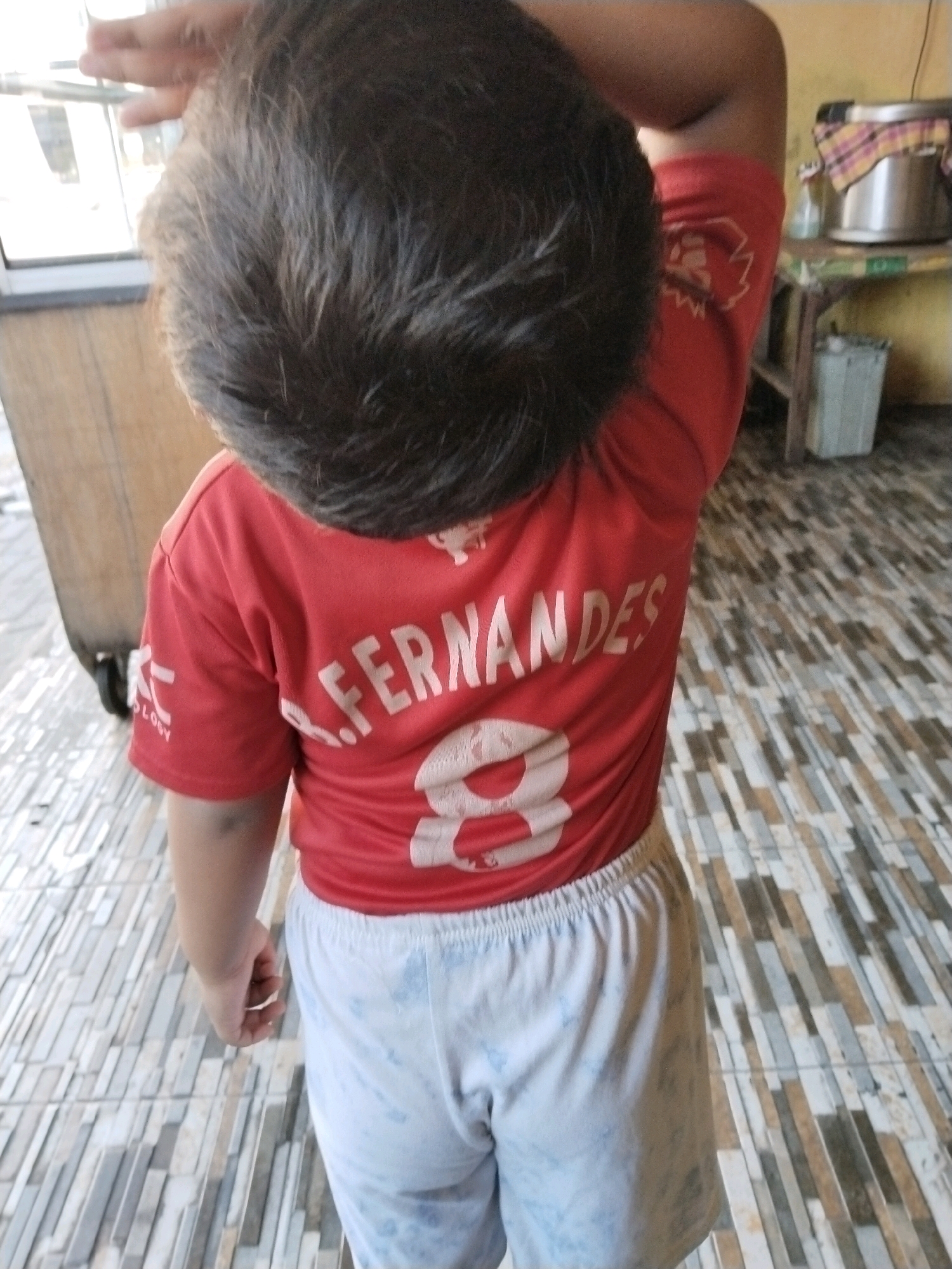 A child, viewed from behind, stands on a tiled floor. The child wears a red t-shirt with white lettering that reads "B. FERNANDES 8". The child also wears light blue patterned shorts. The child's right hand is raised, touching the back of their head. In the background, a metal pot with a checkered cloth is visible on a shelf. To the left, a large wooden object with wheels is partially visible. The setting appears to be outdoors or in a covered outdoor area, possibly in front of a building. The lighting suggests it is daytime. The location is identified as Pepelegi, Indonesia.
