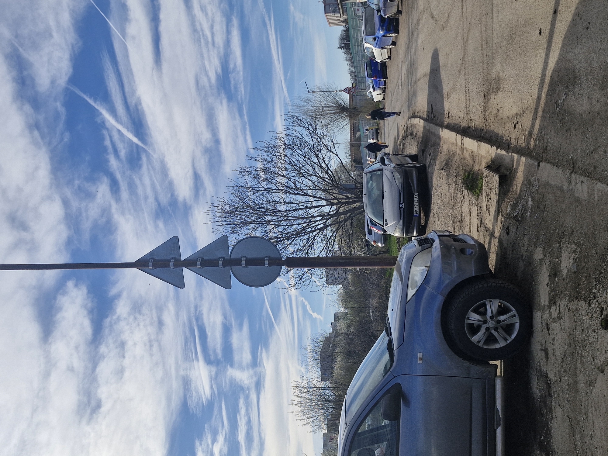 The image depicts a street in Sofia, Bulgaria, with several parked cars. The sky is blue with wispy clouds and visible contrails. A road sign with three direction indicators is present. Two pedestrians walk on the sidewalk. A blue car is parked on the right side of the image.