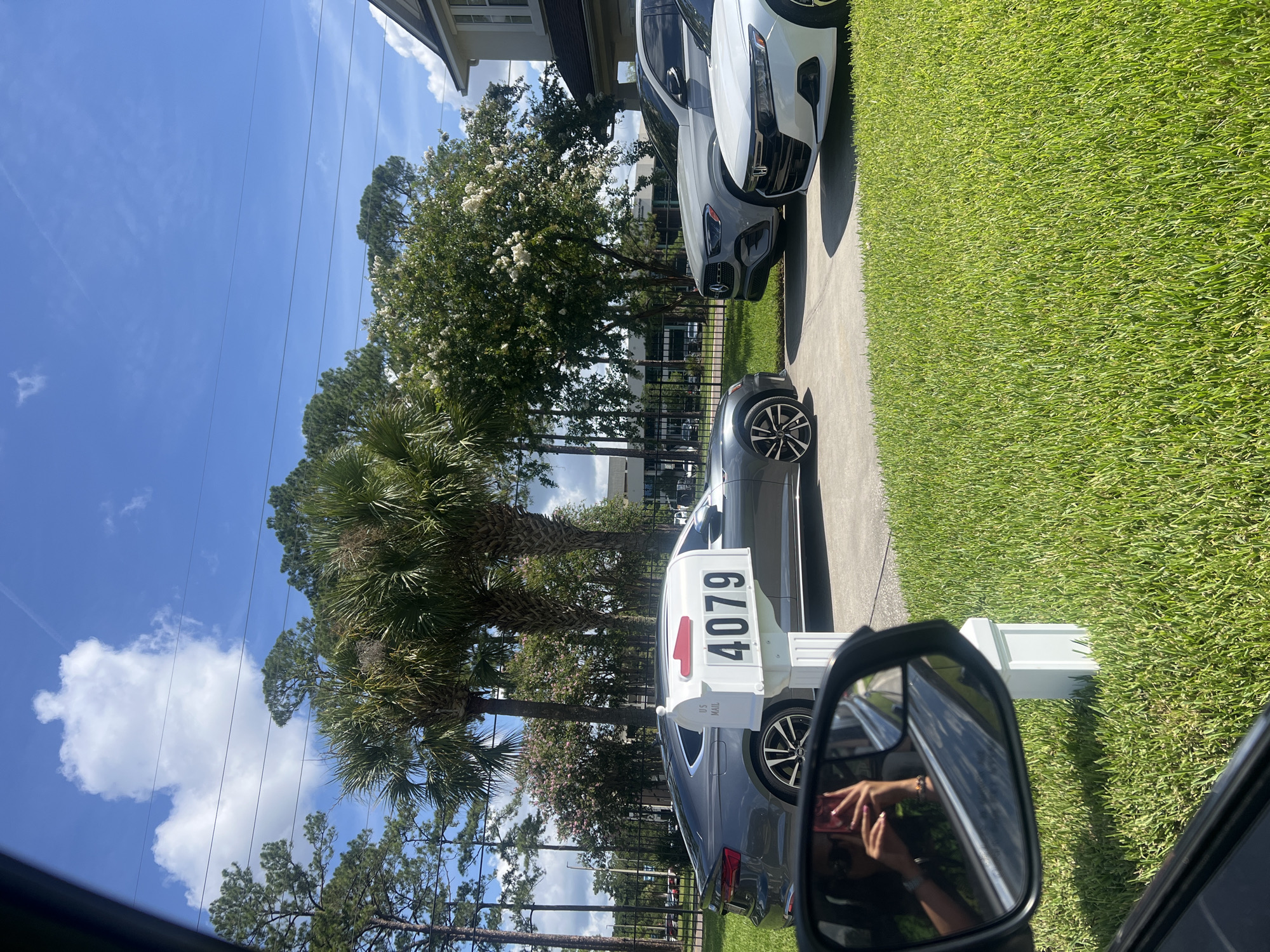 This media file captures a suburban street scene in Jacksonville, USA, under a bright, sunny sky with scattered clouds. The perspective appears to be from inside a car, looking towards the right side of the frame. A prominent white mailbox with the house number "4079" is visible, featuring a red mailbox flag.
Several cars are parked along the driveway, including a white luxury vehicle and a dark-colored sedan. The foreground is dominated by a well-maintained green lawn, indicating a residential area. Trees, including palm trees and flowering trees with white blossoms, provide a lush, natural element to the scene. The presence of power lines suggests an established neighborhood.
In the driver's side mirror, a reflection shows a person holding a pink smartphone, taking a photo or recording a video, suggesting the media file itself is a captured moment of this activity. The overall mood is serene and typical of a sunny day in a suburban environment. The image captures a slice of everyday life, with a focus on the residential details and the natural surroundings. The time of day is clearly daytime, likely late morning or early afternoon, given the bright sunlight and shadows.