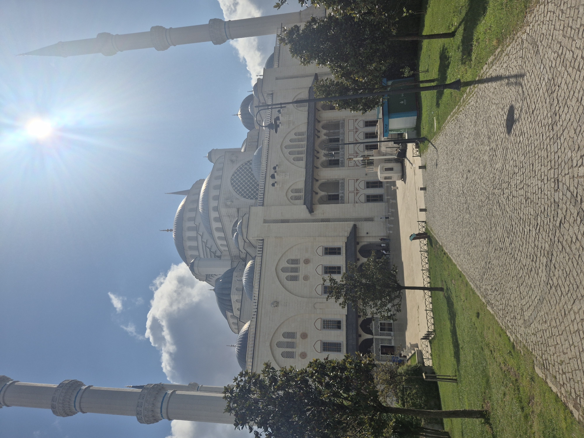 The image shows the exterior of a large, white mosque with multiple domes and tall minarets. The mosque is a prominent structure with intricate architectural details. In the foreground, a cobblestone path leads towards the mosque, bordered by a lush green lawn and several trees. The sky is blue with some scattered clouds, and the sun is shining brightly, casting lens flares and highlighting the facade of the building. There are a few people visible in the distance, walking on the plaza in front of the mosque. A small blue and white booth or kiosk is situated to the right of the path, near some trees. The overall scene depicts a serene and grand religious site bathed in sunlight. This is the Külliye of the Çamlıca Mosque located in Üsküdar, Istanbul, Turkey.