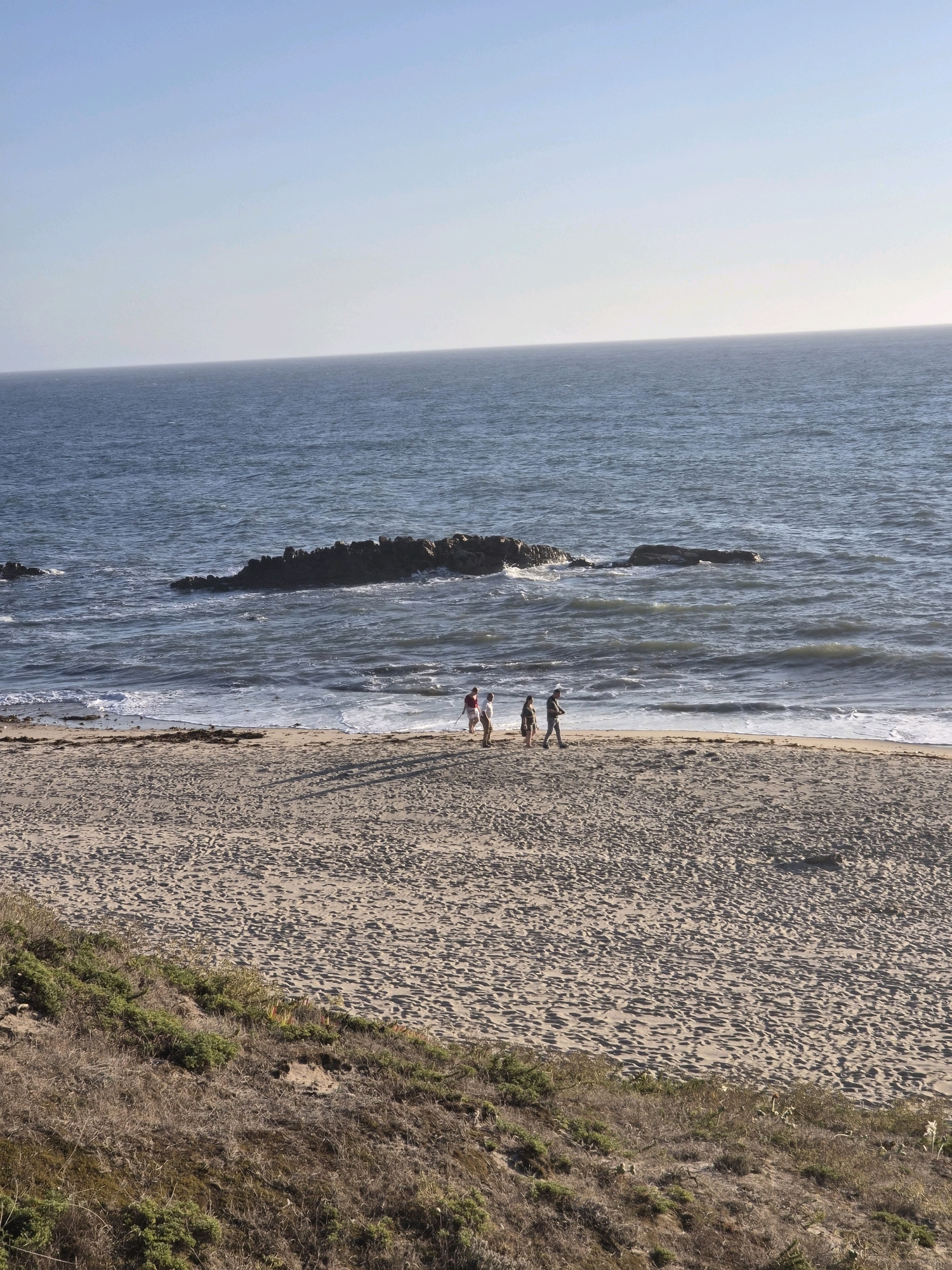 The image shows a wide shot of a sandy beach on a sunny day, with the ocean in the background. The sky is clear and a pale blue. In the middle ground, a group of four people are walking along the shoreline. The ocean has moderate waves, and a rocky outcrop is visible in the water further out. The foreground shows a sandy beach with footprints and some sparse, dry vegetation on a gentle slope. The scene appears to be a natural coastal environment, possibly a park or undeveloped area, as there are no visible buildings or infrastructure associated with a city. While the image depicts a beach, there is no information within the image itself to suggest it is located in Esposende, Portugal.
