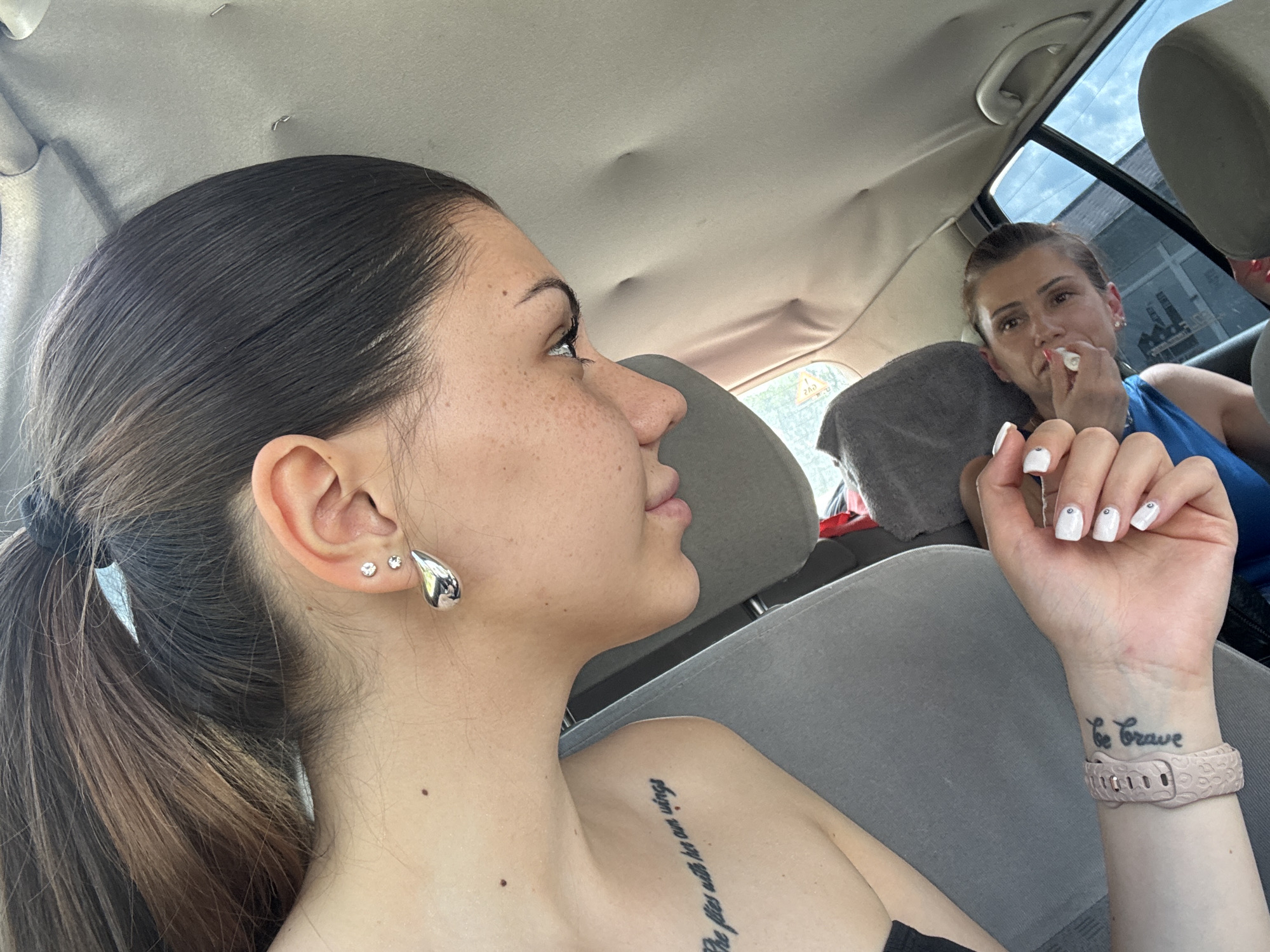 The scene depicts the interior of a vehicle, possibly a car, featuring two adult females. In the foreground, a young woman is seated, angled with her right side towards the viewer and her head tilted downwards. She has dark brown hair, pulled back in a ponytail, with lighter highlights. Her right ear is pierced with two small stud earrings and a larger, polished silver teardrop-shaped earring. Freckles are visible on her cheek and nose. A tattoo reading "She flies with her own wings" is present on her right upper chest/shoulder. Her left hand, with white nail polish and a small rhinestone on the ring finger, holds an indistinct white object. A light pink, textured silicone watch strap is on her left wrist, above which a tattoo reading "Be Brave" is visible. She wears a grey garment over a black undergarment. In the background, another woman with dark hair pulled back is seated in the rear passenger seat, looking forward and appearing to place a small white item into her mouth or wipe her mouth. She wears a blue top. The car's interior features grey fabric seats and a light-colored fabric headliner with several depressions. Through the right rear window, a blurry exterior urban environment is visible under an overcast sky, with faint building structures and a yellow triangular sign with dark markings, potentially Cyrillic, suggesting a road sign. The described scene is located in Sofia, Bulgaria.