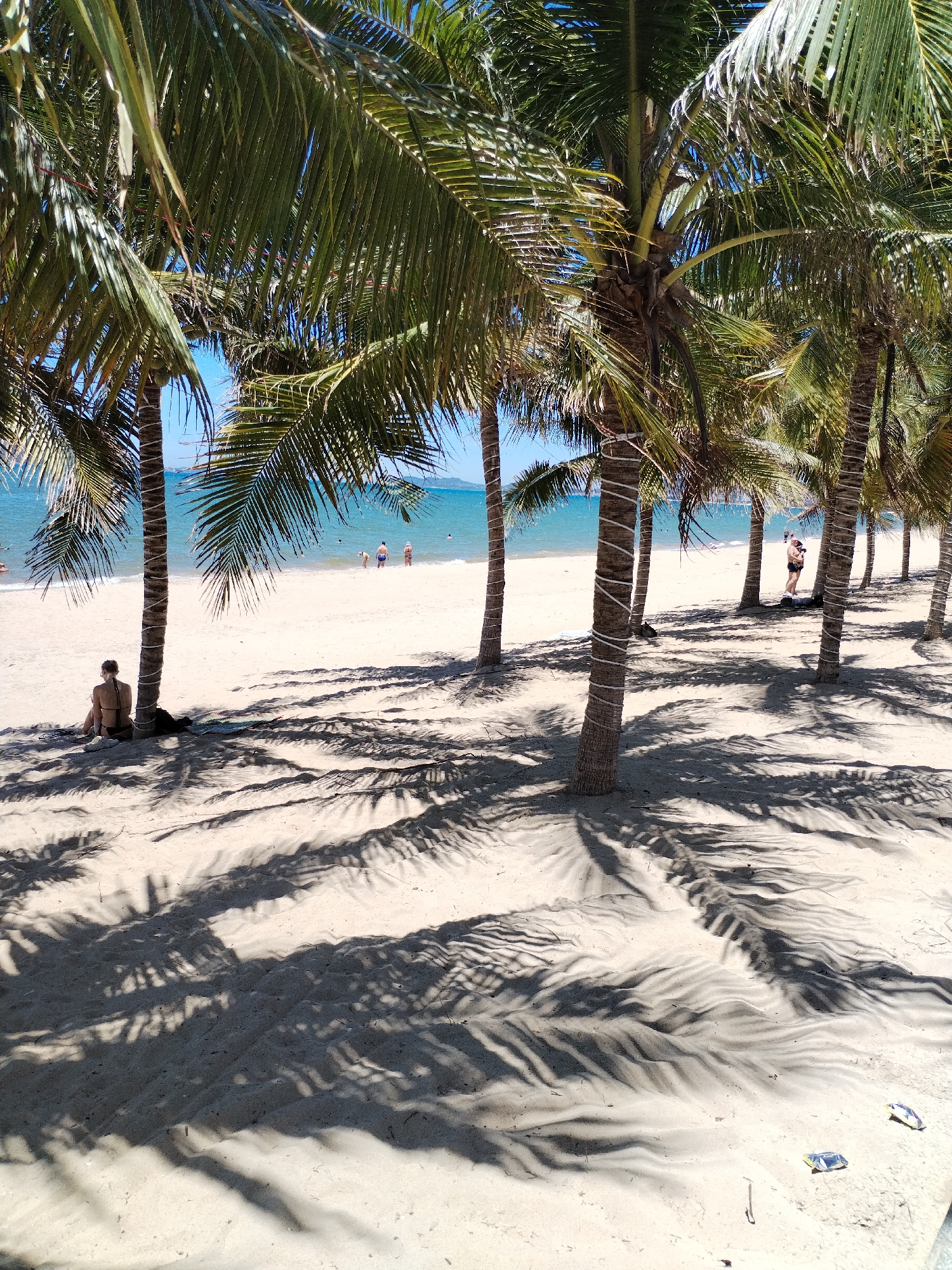 The image captures a beach scene in Nha Trang Ward, Vietnam. Palm trees line the sandy shore, casting shadows across the sand. Several individuals are visible, including a person sitting at the base of a palm tree in the foreground, people swimming in the ocean, and people walking on the beach. The sea is calm with blue water. The light is bright. There are some objects on the sand in the foreground.
