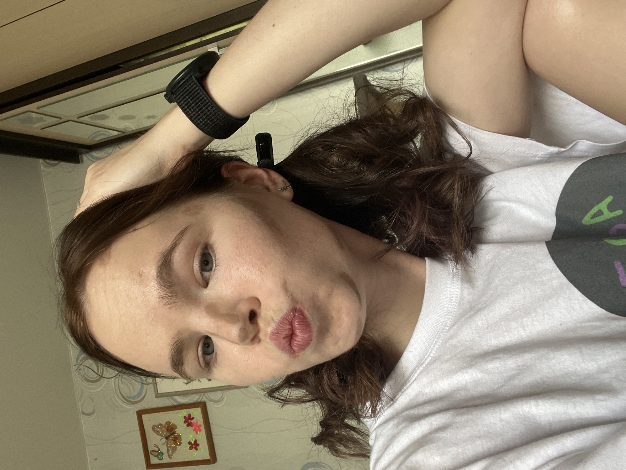 A close-up selfie captures a young woman posing indoors in Chaikovsky, Russia. The image is rotated 90 degrees counter-clockwise, showing her head towards the bottom left and her body extending towards the top right. She has fair skin, dark brown wavy hair, and light-colored eyes, and is making a playful puckered-lip expression.
She is wearing a white short-sleeved t-shirt with a partial dark grey graphic and fragmented purple text on the right side of her chest, which is not legible. On her left wrist, she wears a black digital watch or fitness tracker with a textured strap. A small black hair clip holds back some hair behind her left ear, where a silver hoop earring is visible.
The background suggests a home interior. To the top left, light brown wooden furniture, possibly a cabinet or shelf, is visible. The wall behind her features light-toned wallpaper with a subtle swirling or wavy pattern in shades of blue and grey. Below her chin, a small framed artwork hangs on the wall, showcasing an embroidered butterfly and flowers in orange, pink, and green hues on a light background. The lighting is bright, consistent with a daytime indoor setting. The overall mood of the photo is casual and playful, typical of a personal selfie.
