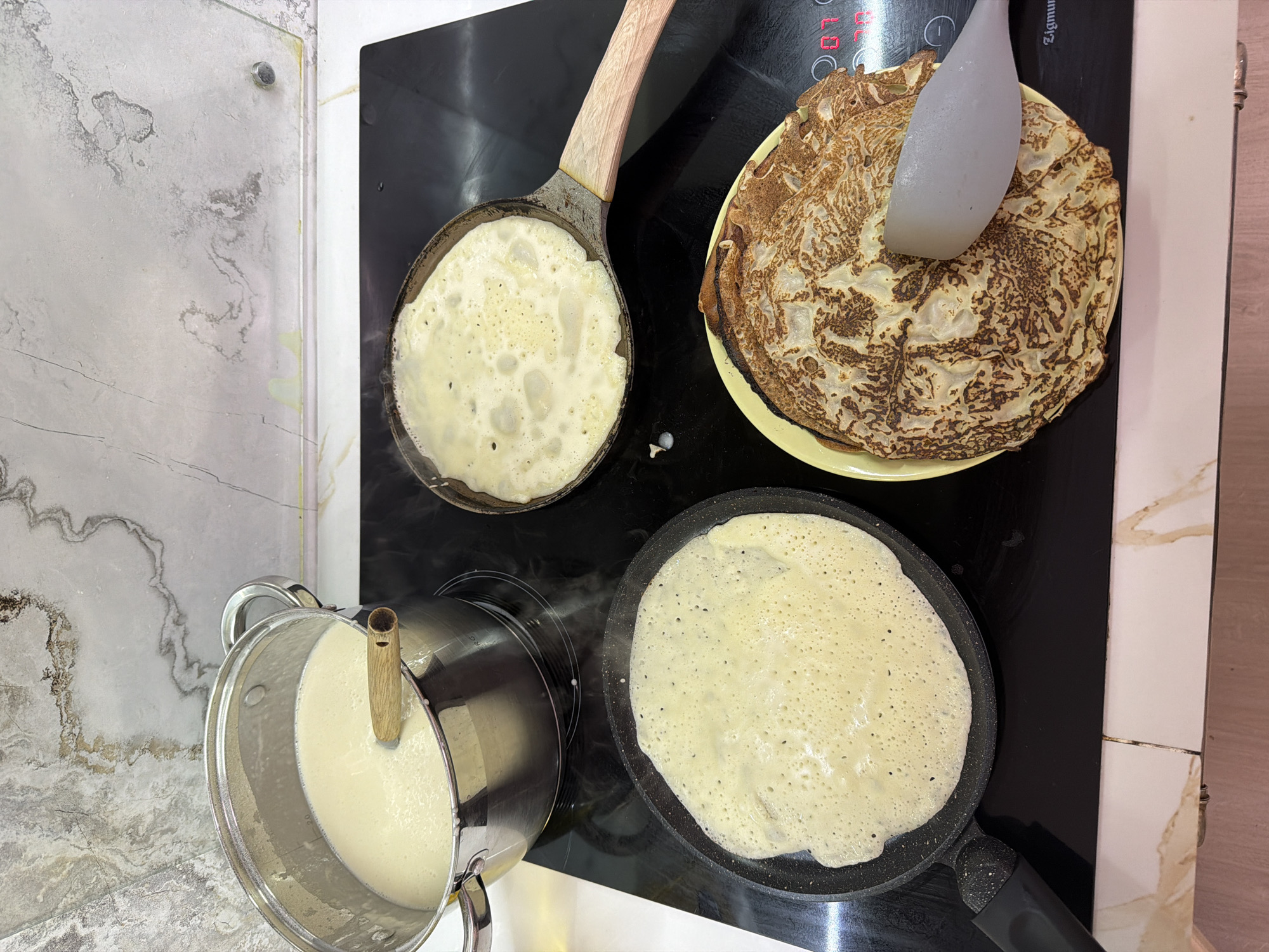 The image shows a domestic kitchen scene with a focus on the preparation of pancakes. A black electric stovetop is in use, with two frying pans containing pancake batter in different stages of cooking. A stack of cooked pancakes sits on a yellow plate, partially covered by a spatula. Next to the stovetop is a metal saucepan containing more pancake batter, with a wooden utensil placed inside. The countertops are a mix of marble-patterned and white tiles. The scene appears to be indoors in a kitchen, likely in a private residence. There are no indications of Sochi, Russia, from visual information.
