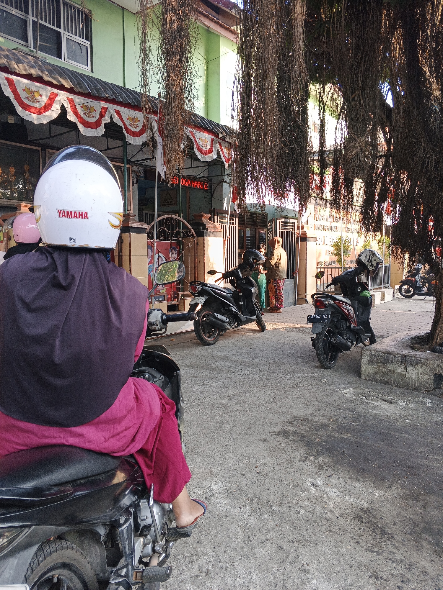 In the city of Krian, Indonesia, the image captures a scene outside a building with a green facade, decorated with red and white banners. In the foreground, a person on a motorcycle, wearing a white Yamaha helmet and a dark purple head covering over a magenta outfit, is viewed from behind. The motorcycle's seat is black, and the person's legs are visible, wearing magenta pants and blue and red flip-flops. To the left, another person on a motorcycle, wearing a pink helmet, is partially visible.
In the midground, several other motorcycles are parked. Two individuals, a woman in a tan hooded jacket and patterned pants, and another person in a green garment, are standing near a gate and a poster featuring children. Behind them, a building with text in Indonesian is visible. Further back, more motorcycles are parked, and another building with a light green wall and barred windows is present. The environment is an outdoor area with a concrete ground, and large, dense tree roots hang down from above, obscuring parts of the scene. The lighting suggests it is daytime, possibly late afternoon, with sunlight filtering through the tree roots.