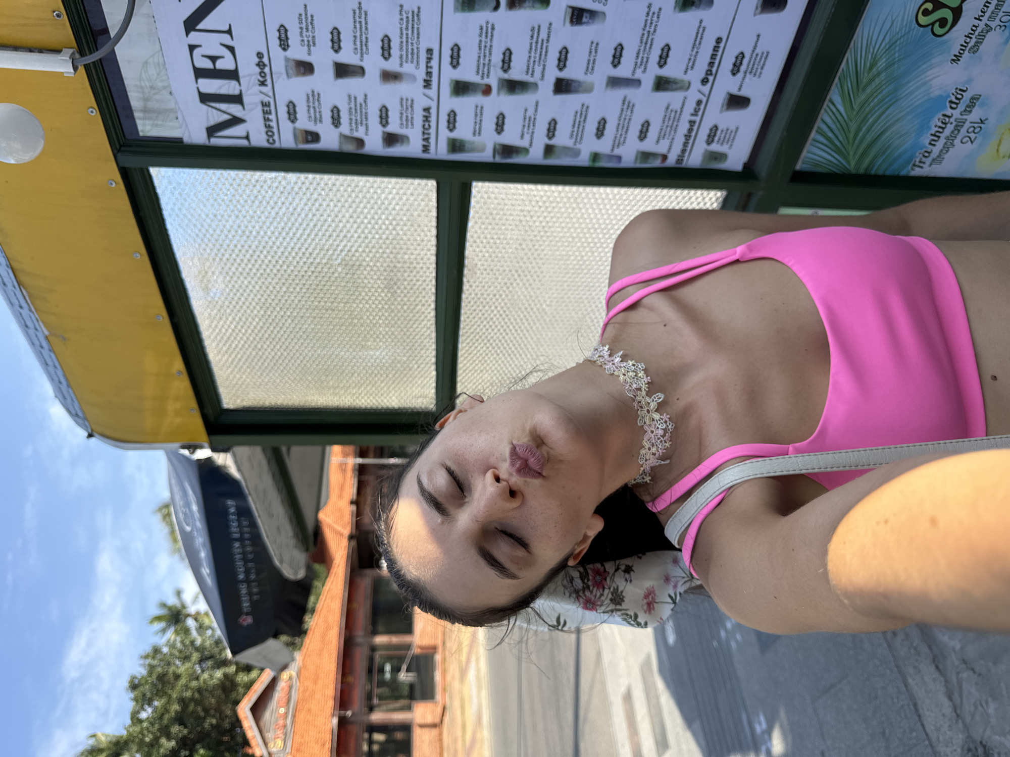 A young woman with her eyes closed and lips puckered leans her head back against a floral patterned fabric attached to a white strap. She wears a bright pink sports bra and a delicate white and pink beaded choker. Sunlight casts a shadow on her arm. Behind her, a glass-fronted kiosk displays a menu with prices in Vietnamese and Russian, featuring coffees, matchas, and blended ice drinks. The menu is illuminated by light bulbs. To the left, a building with an orange tiled roof and wooden accents is visible, along with palm trees and a black umbrella with white text. The scene is set outdoors on a sunny day.
