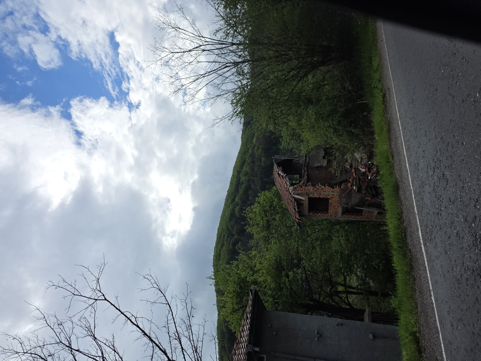 The image shows a ruined brick building with a collapsed roof, located near a road and a green, forested hillside. A grey building is visible in the lower part of the image. The sky is partly cloudy. The location is Gintsi, Bulgaria.