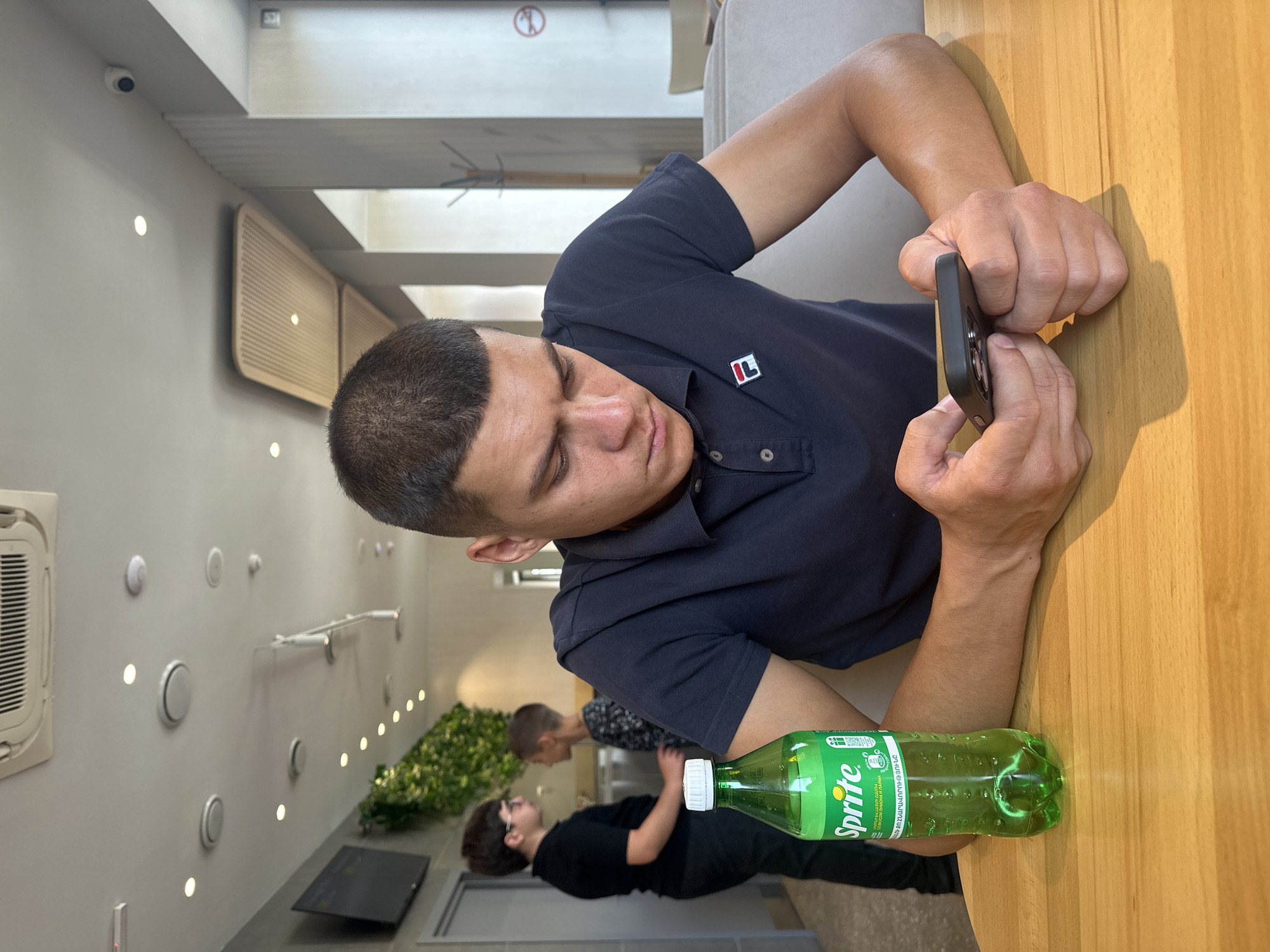 A young man with a buzz cut, wearing a dark blue polo shirt with a Fila logo on the left chest, is seated at a light brown wooden table, engrossed in his smartphone. His hands are clasped around the phone, and he appears to be typing. To his left on the table is a clear green bottle of Sprite with a white cap and label. In the background, two young individuals are standing near a wall with artificial greenery. The environment appears to be an indoor public space, possibly a cafe or restaurant, with a tiled ceiling and air conditioning unit visible. The overall lighting suggests it is daytime. The location is specified as Gyumri, Armenia.