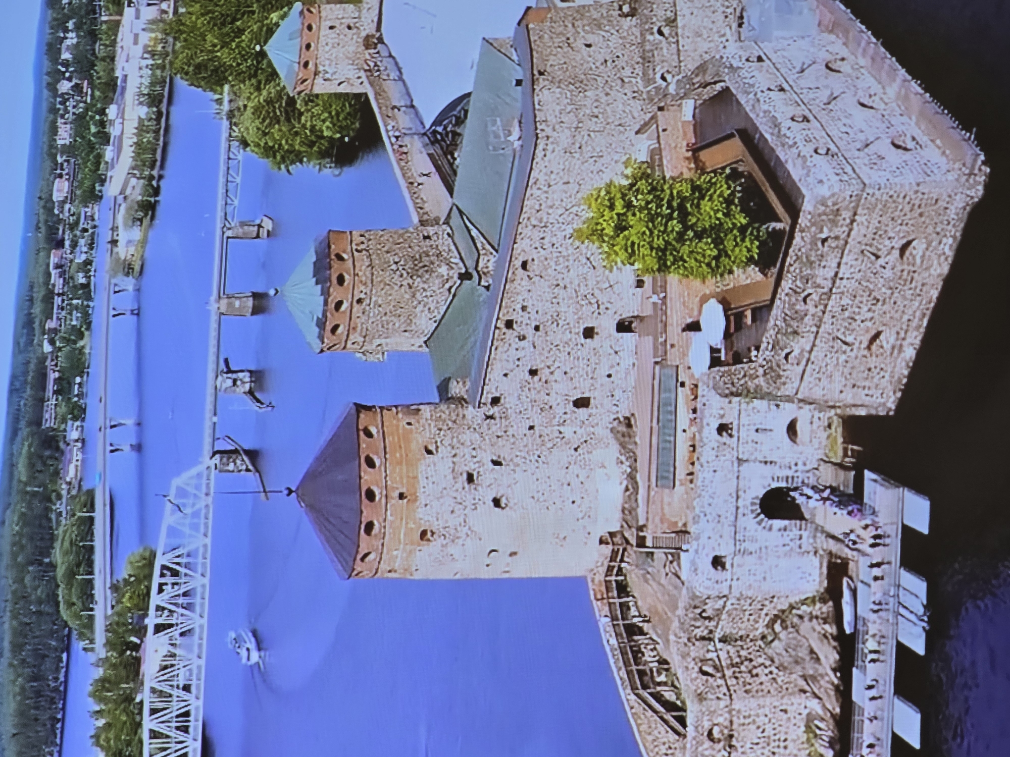 This is an aerial view of Olavinlinna Castle, located in Savonlinna, Finland, not Helsinki. The castle is situated on a rocky island in the Saimaa lake system. The stone fortress features multiple towers with conical, dark-colored roofs. A green canopy is visible on one of the lower structures of the castle. In the foreground, a wooden walkway leads to the castle entrance, where a group of people are visible. Several other individuals are present on a floating dock structure attached to the walkway. A small white boat is moored to the dock. In the background, a large white truss bridge spans a body of water, with a white boat passing underneath it. Another bridge with concrete piers is visible further in the distance, leading towards a town with buildings nestled amongst green trees. The water is a deep blue, reflecting the sky, and the overall scene is bathed in daylight.