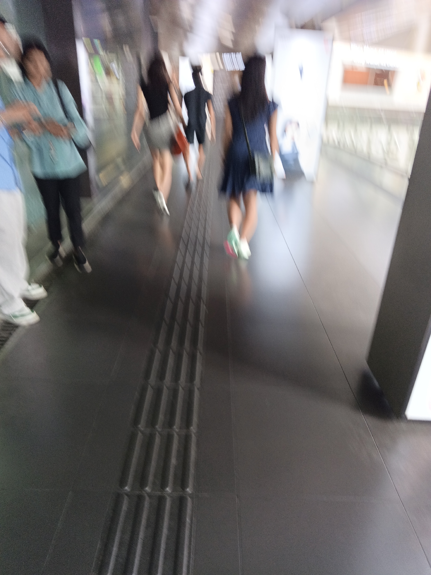 The image captures a blurred scene of people walking in an indoor environment, possibly a concourse or walkway in Kuala Lumpur, Malaysia. In the foreground, a person in light-colored pants and white shoes is partially visible on the left. To their right, a person in a light blue top and black pants is walking away from the camera. Further back, several other individuals are walking in the same direction. One person wears a black top and a grey skirt, and another is in a black t-shirt and shorts. A woman in a dark blue dress with a black bag slung over her shoulder is also visible. The floor is dark gray and features a textured tactile paving strip running along the right side of the walkway, indicating a public space possibly designed for accessibility. The background is indistinct due to motion blur, showing architectural elements and lighting, suggesting an urban indoor setting.