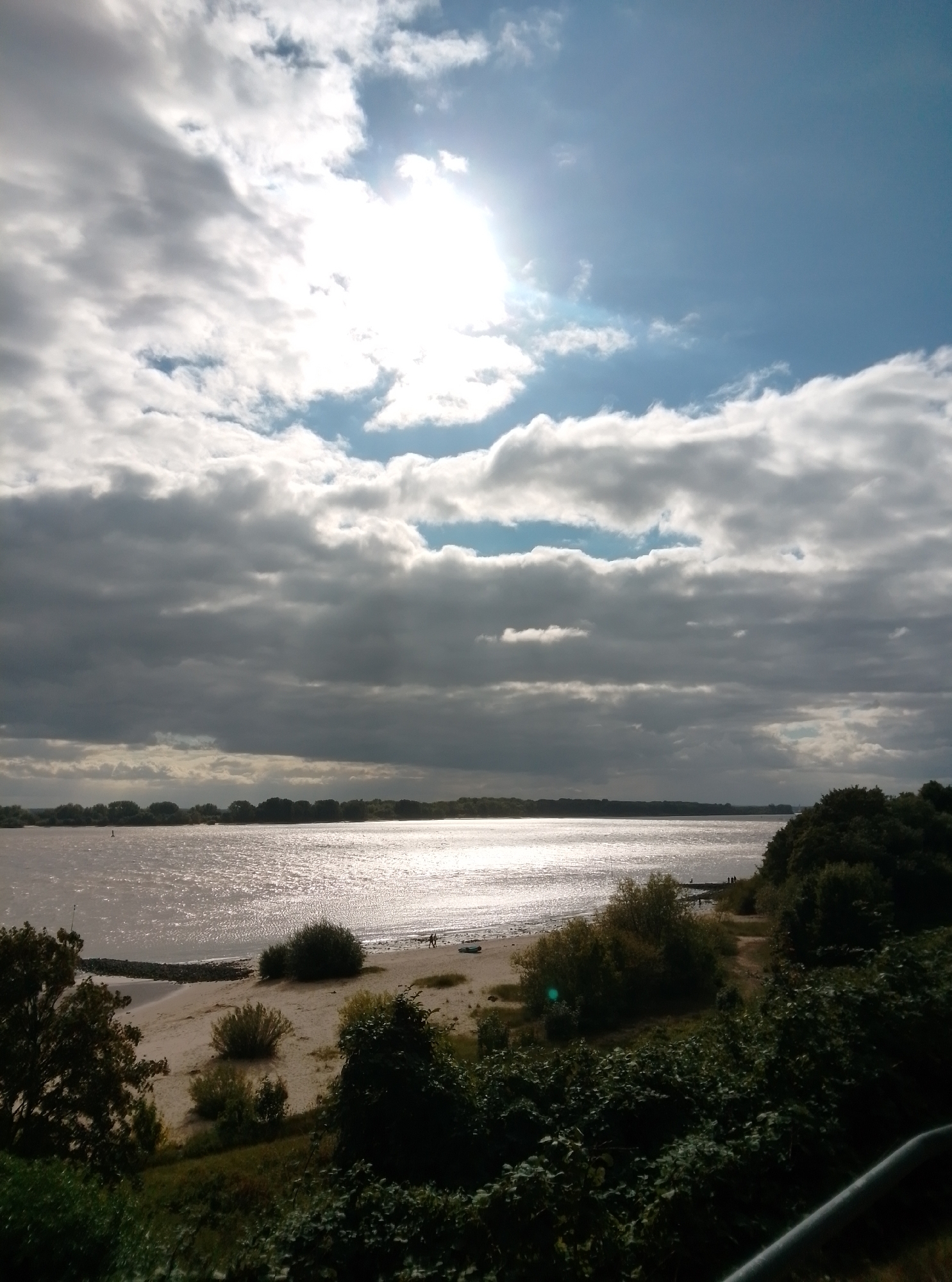 The image depicts a wide, calm body of water, likely the Elbe River, with a sandy beach in the foreground. The sky is a mix of bright blue and heavy grey clouds, with the sun partially obscured by clouds, casting a shimmering reflection on the water's surface.
On the beach, two individuals are visible walking near the water's edge. A small boat is also present on the sand. The coastline is lined with a dense band of trees, forming a dark silhouette against the lighter hues of the water and sky. In the distance, a hazy treeline suggests more land across the river.
The foreground of the image is dominated by lush green foliage and bushes, with a dark, possibly metallic railing visible at the bottom right corner, suggesting a viewpoint from an elevated position. The overall scene evokes a sense of a peaceful, late afternoon or early evening at a waterfront location in Wedel, Germany.