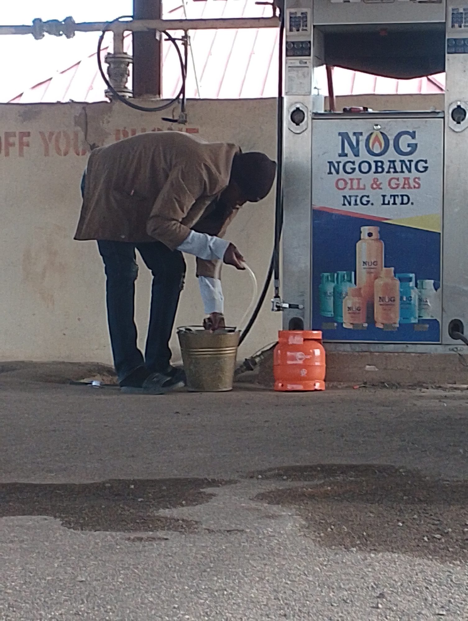 A person is seen at a fuel station in Kwang, Nigeria. The individual, wearing a brown jacket and black pants, is bent over a golden bucket, appearing to pour liquid from a white hose into it. Next to the bucket, an orange gas cylinder is visible. The fuel pump behind the person displays the logo "NOG NOGOBANG OIL & GAS NIG. LTD." The ground is asphalt with some dark stains, possibly from spilled fuel. The background reveals a structure with a red roof and a pale wall with faint lettering.