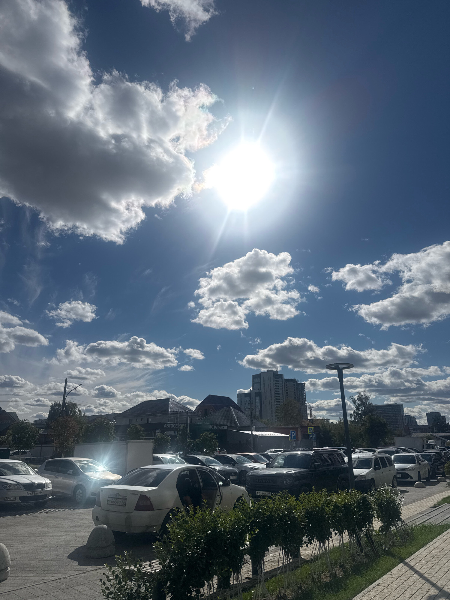 The image depicts a daytime scene in a city, likely an outdoor parking lot. The sky is bright blue with scattered white and grey clouds. The sun is prominently visible in the upper portion of the frame, creating lens flare. In the foreground, several cars are parked. A white sedan is in the center, with its driver's side door open and a person partially visible inside. Other cars, including a black SUV and various sedans and hatchbacks, are parked in rows. Behind the parking lot, low-rise buildings with pitched roofs, some appearing to be commercial structures with signage, are visible. Further in the background, taller residential or office buildings can be seen. A streetlamp stands near the center of the image. Greenery, including bushes and trees, is present in the foreground and between parking rows. The overall impression is that of a typical urban parking area on a partly cloudy day. The location is identified as Novosibirsk, Russia.