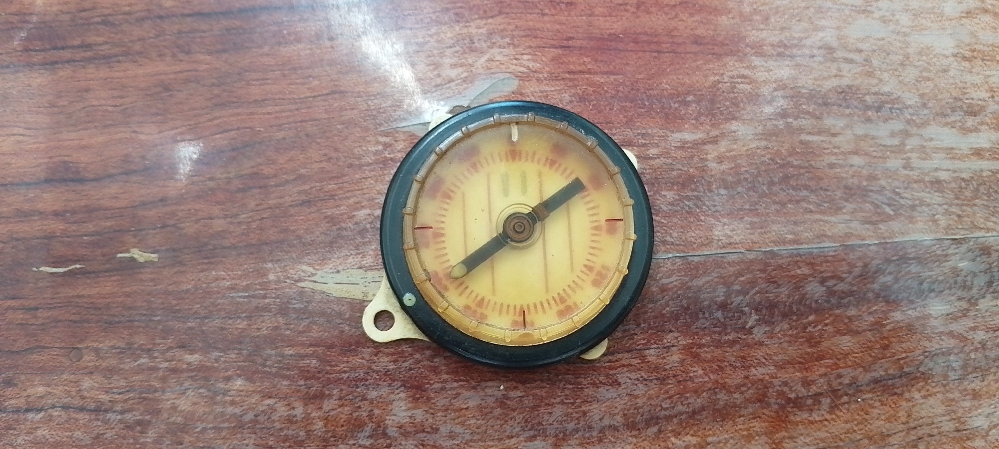 A round, vintage gauge with a black rim and a yellow face is centrally positioned on a textured, dark brown wooden surface. The gauge has a black needle pointing towards the upper left quadrant, with tick marks and some red markings on the dial. There are two mounting tabs attached to the bottom right of the gauge. The wooden surface appears to be a table or countertop with visible grain patterns and some light wear. The image was captured in Cherkasy, Ukraine.