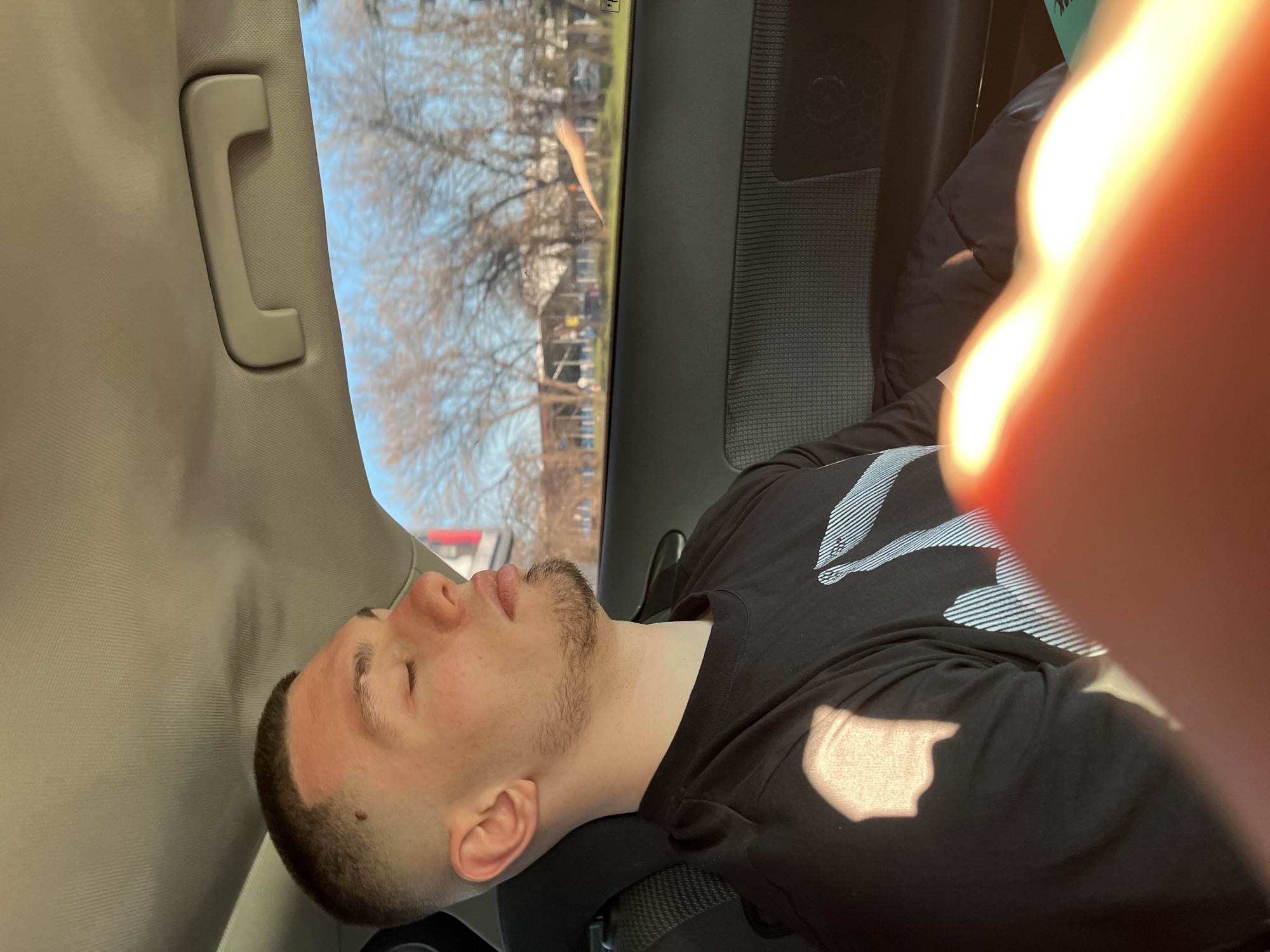 A man is reclining in what appears to be the passenger seat of a car. He has short brown hair, a light beard, and is wearing a black t-shirt with a white graphic design. He is facing the left side of the image with his eyes closed. The interior of the car is visible, including a passenger side grab handle and a window showing trees and buildings outside. Part of a hand is in the foreground, out of focus and obscuring some of the scene. The image does not contain the location of Burgas, Bulgaria.