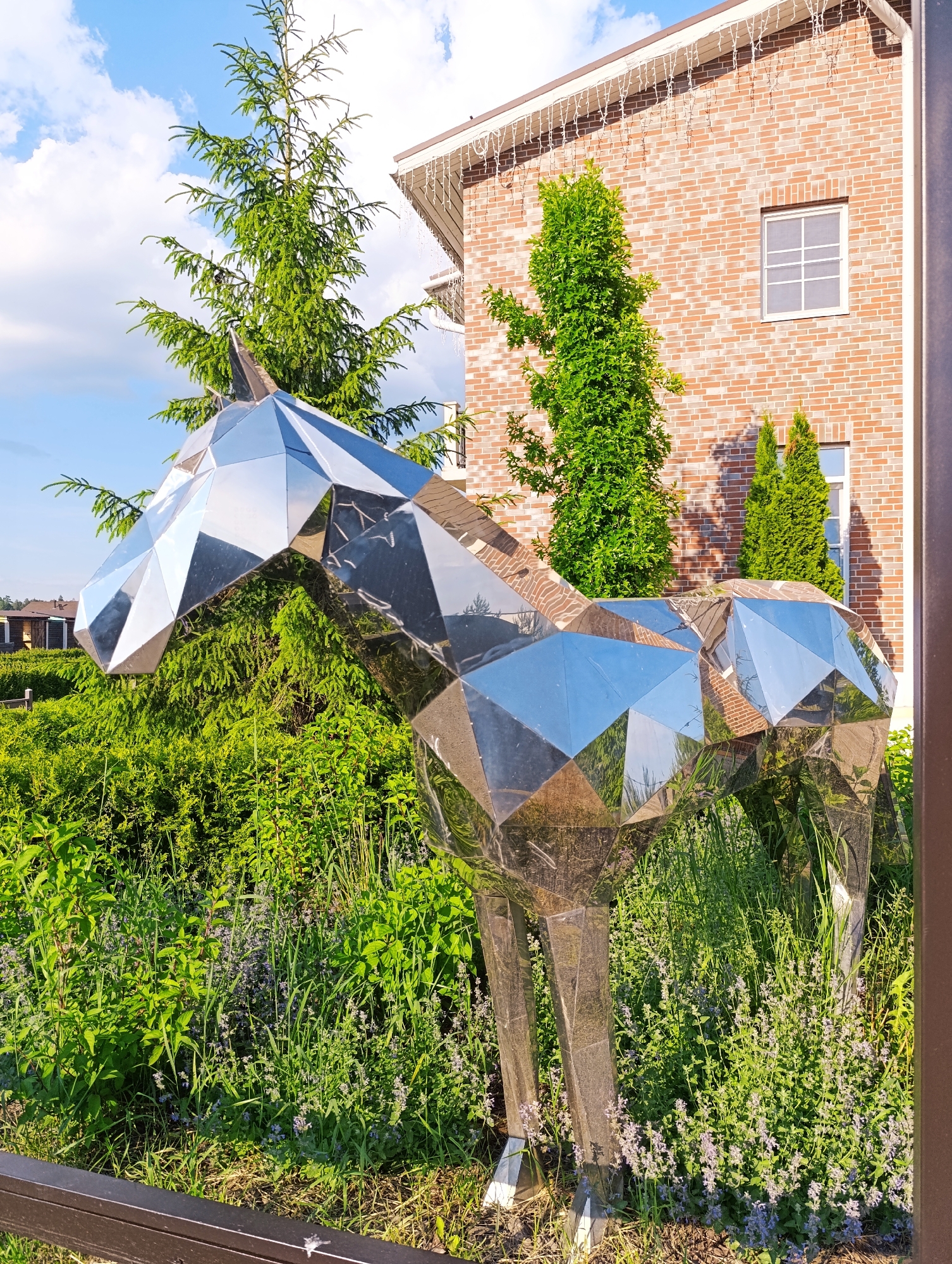A modern, geometric sculpture of a horse made of polished metal stands in a garden setting. The sculpture is highly reflective, mirroring its surroundings. Behind the sculpture, a brick building with white-framed windows and white icicle-like decorations along the roofline is visible. Various green trees and lush vegetation, including plants with small purple flowers, surround the sculpture. The sky is blue with scattered clouds. The scene appears to be outdoors, possibly in a residential or landscaped area. The specific location is a street in Ivanovskoe, Russia, as indicated by a partial sign visible in the image.