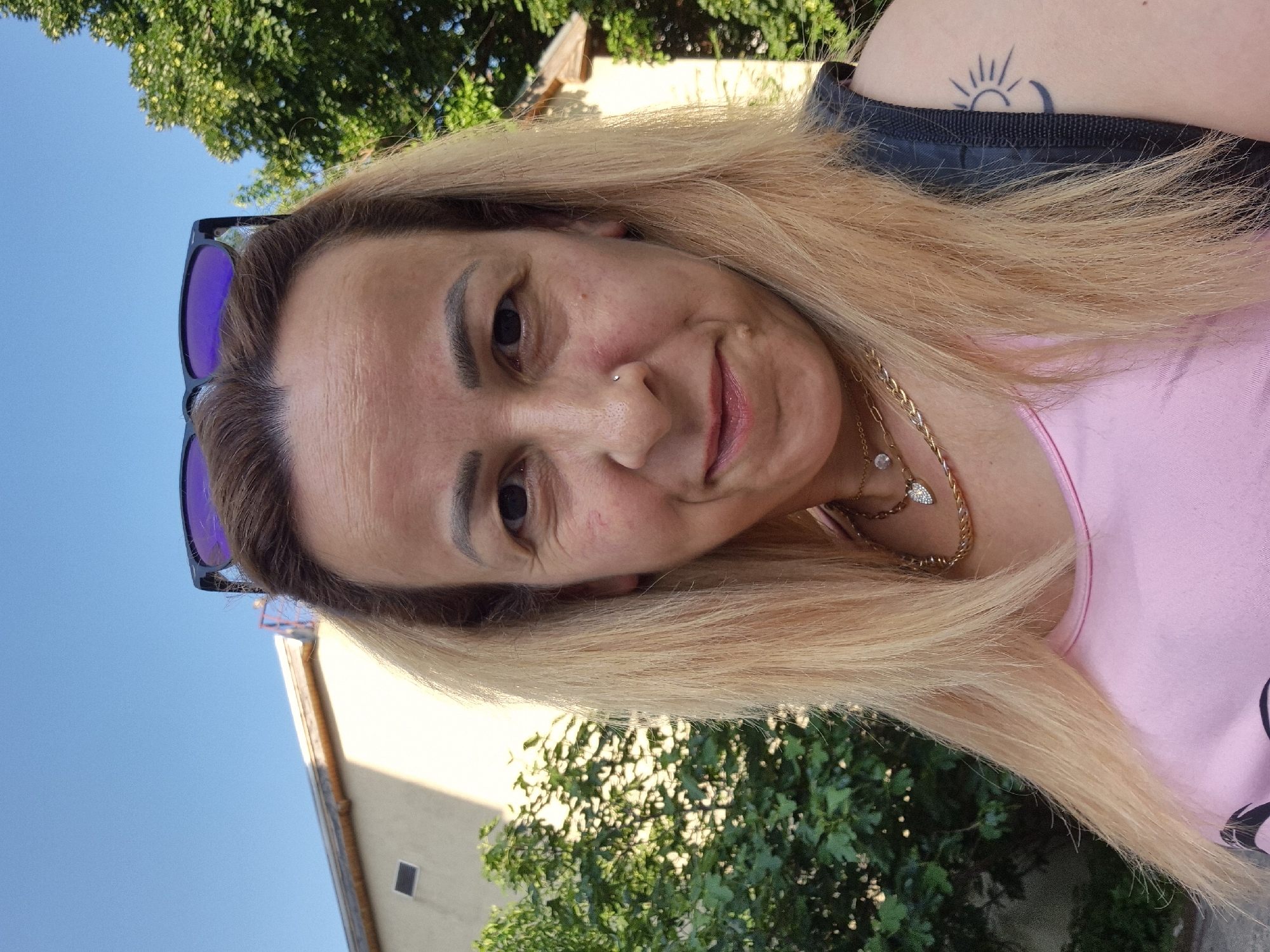 A female individual is depicted in a head-and-shoulder portrait taken outdoors under a clear blue sky. She has shoulder-length, light-blonde hair with darker roots, and is wearing sunglasses with purple reflective lenses pushed up on her head. Her face shows visible forehead wrinkles and a small silver stud piercing on her left nostril. She has a slight smile and is looking directly at the camera. She is wearing a pink top and multiple gold-toned necklaces, including a thicker chain and a thinner chain with a heart-shaped pendant and another circular clear gem pendant. A black strap, consistent with a backpack or bag, is visible over her right shoulder, where a tattoo of a stylized sun and crescent moon is also present on her upper arm. The background features a light-colored building with a roofline and a dark rectangular vent on the left, partially obscured by green bushes. A dense green tree is visible on the right side of the background. The lighting indicates a bright, sunny day. The precise location within the city of Unknown, Unknown, is not discernible.