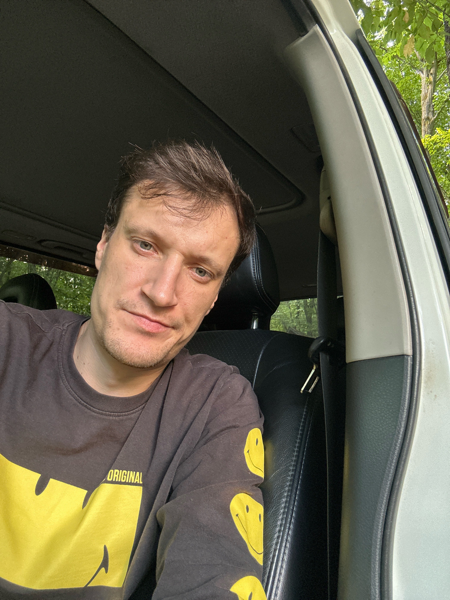 A man with light skin and brown hair is sitting in the driver's seat of a car, looking at the camera. He is wearing a dark gray long-sleeved shirt with a large yellow smiley face graphic and the word "ORIGINAL" printed on it. The car's interior is visible, including the black leather seat and a portion of the car's headliner. Through the open car door, lush green trees and foliage are visible, suggesting an outdoor setting. The lighting indicates it is daytime, likely with sunlight filtering through the trees. The scene appears to be in a rural or wooded area, possibly near Shatava, Ukraine, given the context.