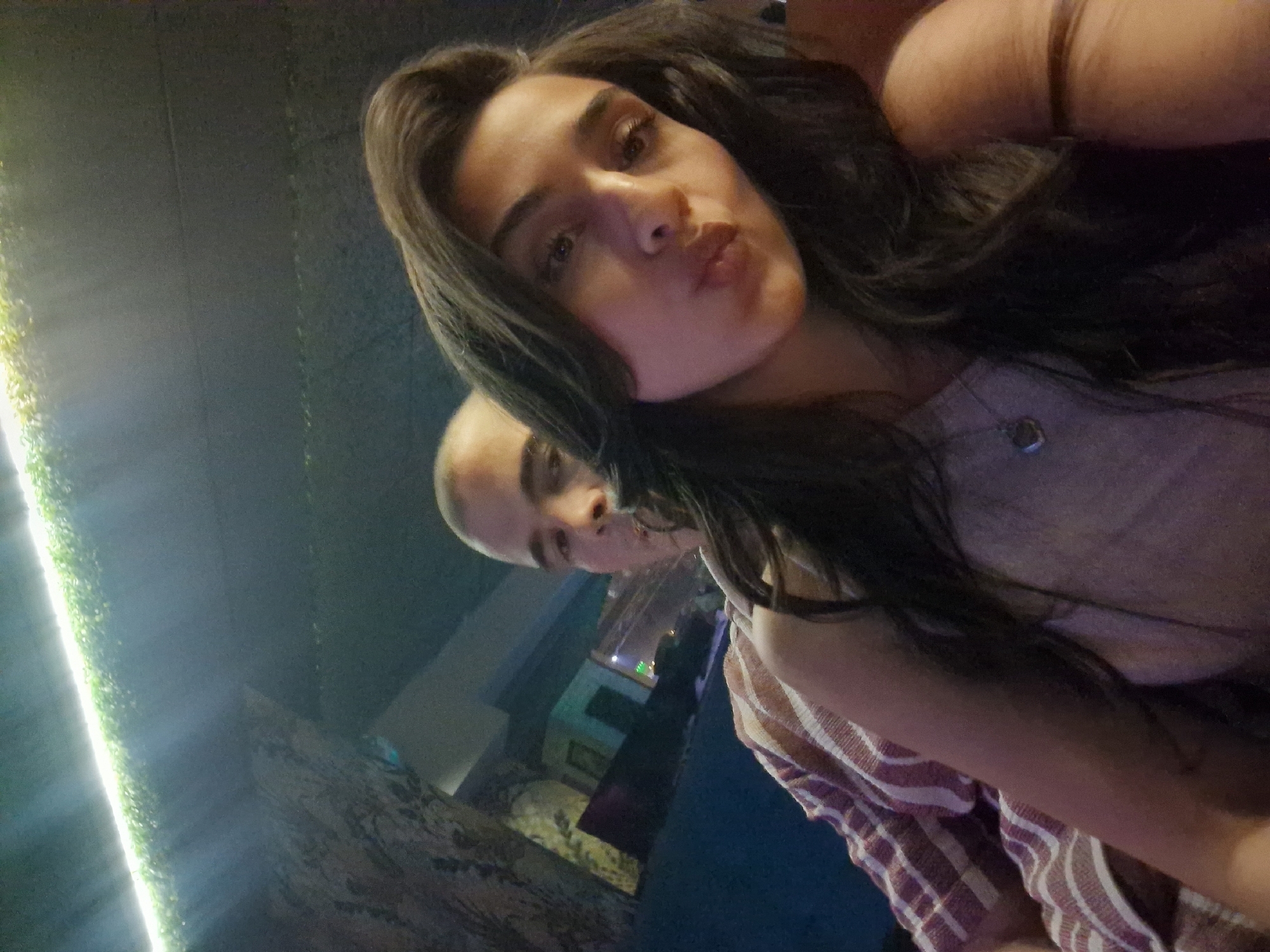 A close-up, selfie-style photograph shows a young woman in the foreground, her face turned towards the camera with her lips pursed. She has long, dark, wavy hair and is wearing a light-colored top with a delicate necklace. Behind her, a bald man is partially visible, his head and shoulder taking up the left side of the frame. He is wearing a striped shirt. The background is dark and appears to be an indoor environment with textured walls and a strip of bright, green-tinted lights running along the top. The overall lighting is dim and atmospheric.