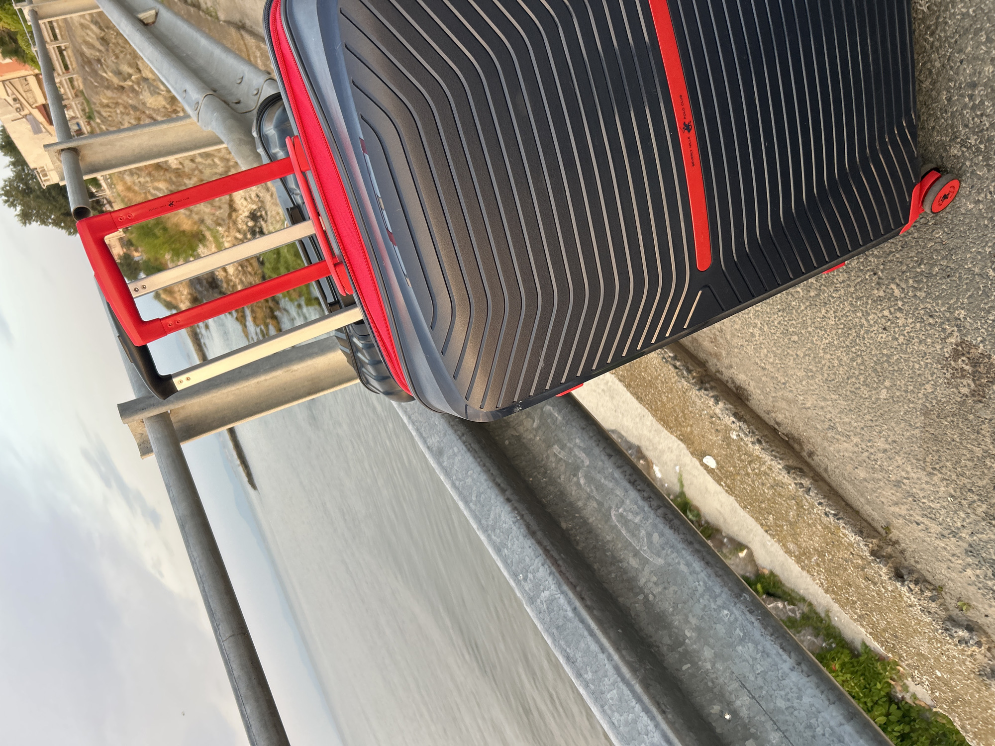 A black suitcase with red accents and a retractable red handle stands on a concrete surface next to a metal guardrail. Beyond the guardrail, the calm water of the sea stretches out under a cloudy sky. In the background, buildings and trees are visible along the coastline. The scene appears to be a scenic spot in Ravda, Bulgaria, possibly near the sea or a promenade.