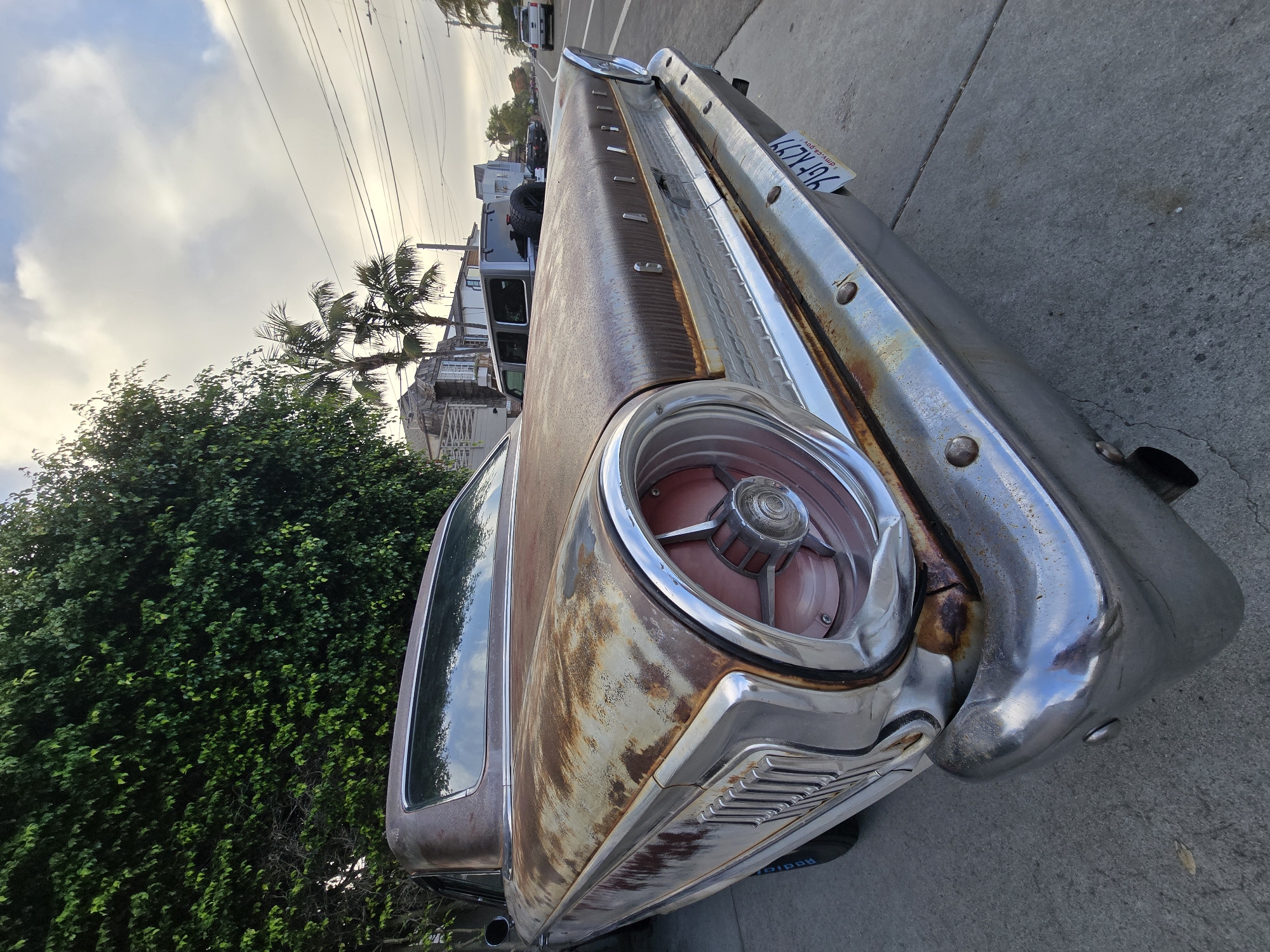 An aged, rusty classic car, possibly a 1950s or 60s Ford, is parked on the side of a street in Encinitas, California. The car is brown with significant rust patches and a peeling paint job. Its chrome bumper is still somewhat shiny, reflecting the sky. The rear taillight is a prominent circular design with a chrome bezel and red lenses. A California license plate reading "YGF KLY9" is attached to the bumper. The car is parked next to a sidewalk and a dense green hedge. In the background, there are palm trees and buildings, suggesting a residential or mixed-use area. The sky is partly cloudy with the sun low on the horizon, indicating it might be early morning or late afternoon. Other vehicles, including a modern SUV and pickup truck, are visible further down the street, along with power lines.