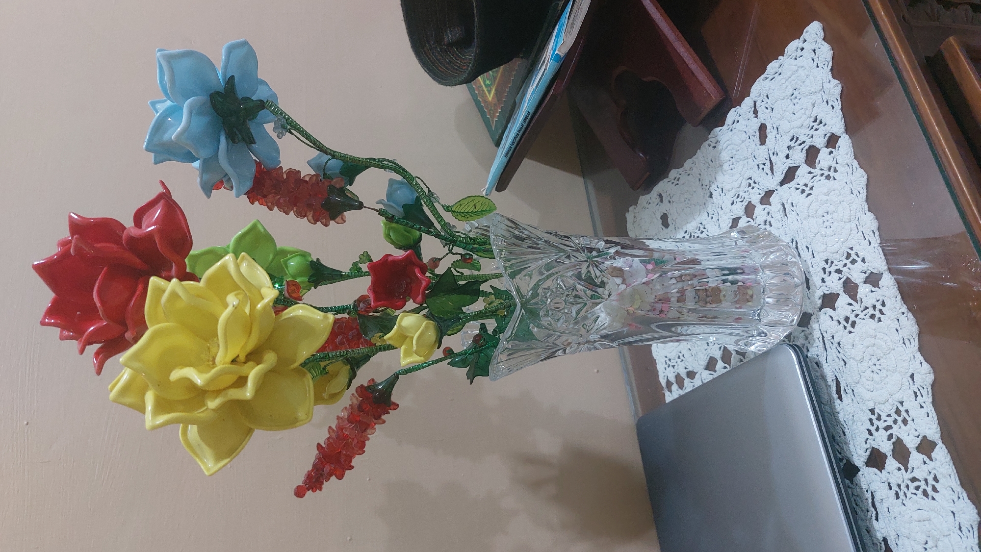 A close-up, eye-level shot captures a decorative arrangement of artificial flowers in a crystal vase, placed on a wooden table. The flowers, in vibrant red, yellow, and blue, are meticulously crafted and arranged on slender green stems, creating a colorful bouquet. The vase, made of clear, faceted crystal, showcases intricate patterns and reflects the light, adding to the visual appeal.
Beside the vase, a silver laptop rests on a white crocheted doily, suggesting a home or office setting. To the right, a wooden stand holds an open book, possibly religious in nature, with a dark, woven cap placed on top. The background consists of a plain, light-colored wall, providing a simple backdrop that emphasizes the floral arrangement and the surrounding objects.
The lighting is soft and diffused, creating subtle shadows that add depth and dimension to the scene. The overall atmosphere is one of domestic tranquility, with the arrangement of objects suggesting a thoughtful and personalized space. While the image provides a detailed view of the objects, it does not offer any clues about the specific location in the city of Medan, Indonesia, beyond what can be inferred from the general style of the objects and the interior setting.