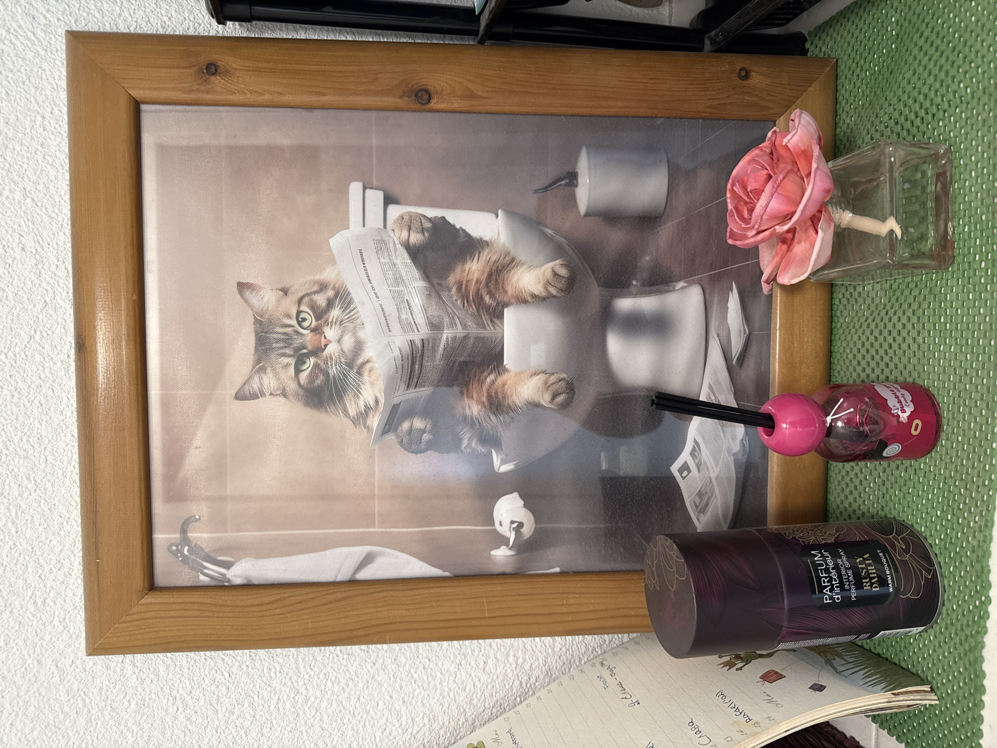 An indoor scene displays a vertically oriented framed picture positioned centrally on a green, textured mat. The picture's wooden frame is light brown with visible wood grain and corner joint details. The artwork within the frame depicts a long-haired, tabby-patterned cat with green eyes, seated on a white toilet. The cat holds an open newspaper, appearing to read it, with its front paws holding the paper and its hind legs extended over the front of the toilet bowl. Behind the toilet, a white toilet brush holder is visible on a dark floor. To the left of the toilet, a white towel hangs from a dark hook, above a white toilet paper holder with a roll. The background within the picture shows light brown tiled walls.
On the green mat in front of the framed picture, from left to right, are three distinct objects. On the far left is a dark maroon cylindrical spray can with a decorative pattern, labeled "PARFUM d'intérieur / INTERIOR PERFUME SPRAY / RUSTY DAHLIA / WARM BOUQUET." Next to it is a small, pink, cherry-shaped air freshener bottle containing a reddish liquid, with several black diffuser sticks protruding from its top. A partial label on the bottle indicates "Bubble G... Candy." To the right, a clear, rectangular glass vase holds a single, artificial pink rose with multiple petals.
Beyond the frame, to its left, a white wall with a textured, stucco-like finish is visible. Partially obscured by the perfume spray can, an open notebook or planner with handwritten text, dates (e.g., 13, 15, 16), and some simple drawings (e.g., a kite) rests against the wall. To the right of the frame, a dark, vertical structure, possibly part of a shelf or furniture, is partially visible. The green mat extends across the foreground surface.