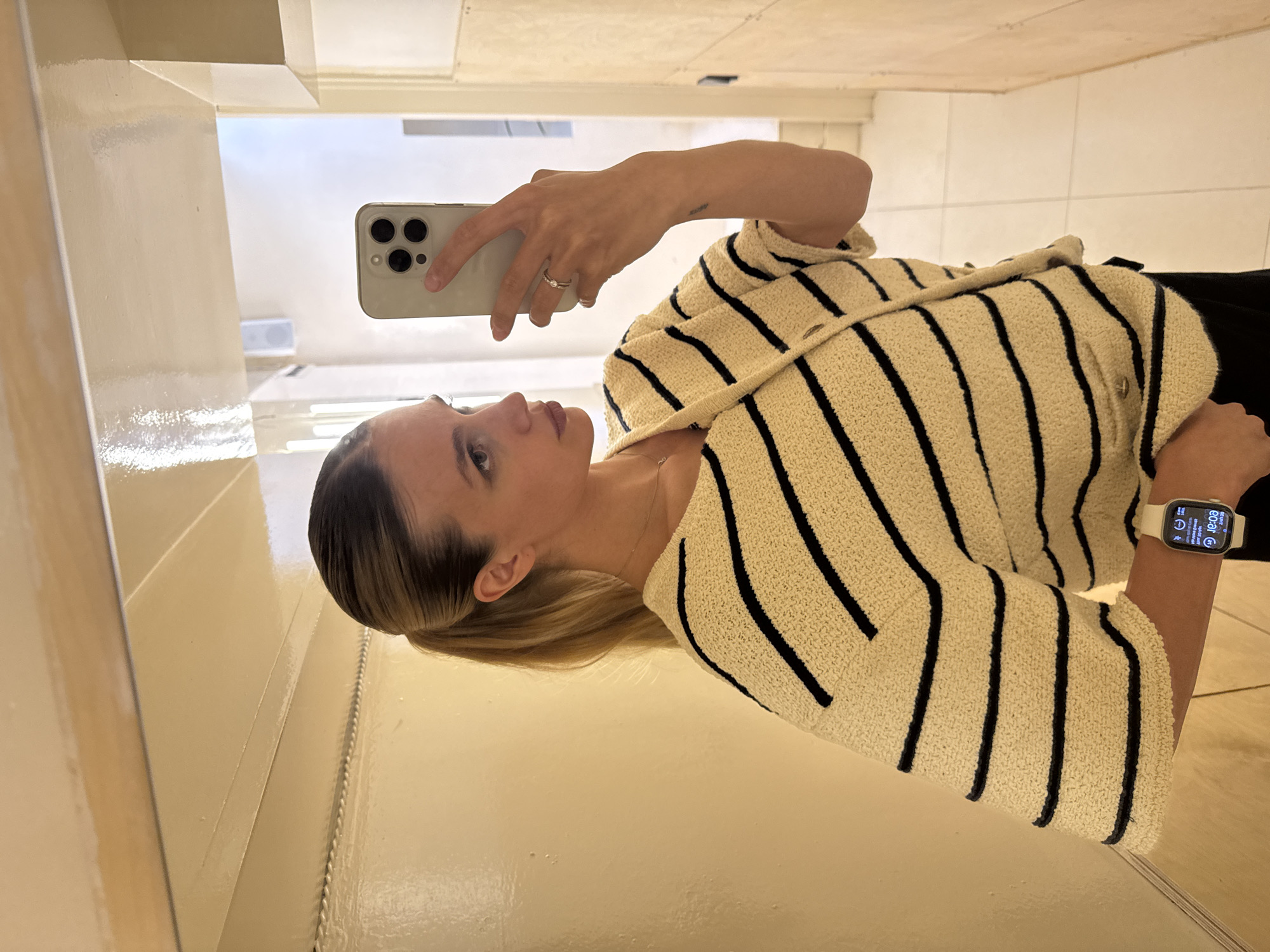 A woman is captured in a mirror selfie, with the image displayed rotated 90 degrees counter-clockwise. She holds a light-colored iPhone, equipped with a triple camera system, with her left hand to take the picture. Her face is shown in profile, looking slightly upwards and to the left, with a neutral expression. Her light brown or dark blonde hair is pulled back, revealing a simple necklace and small hoop earrings.
She is wearing a textured cream or beige cardigan with prominent black horizontal stripes, layered over what appears to be dark-colored clothing like a top or trousers. On the ring finger of her left hand, a silver ring with a clear stone is visible. A small, indistinct tattoo is on her left forearm. On her right wrist, she wears a white-banded smartwatch, its screen displaying "00:01 AM," "G-VENT," and other small digital icons, indicating an event notification.
The setting is an indoor space with highly reflective, light-colored walls or surfaces acting as the mirror. In the background reflection, parts of a ceiling with light fixtures are visible, suggesting a modern interior. The context provided for the location is Tbilisi, Georgia.