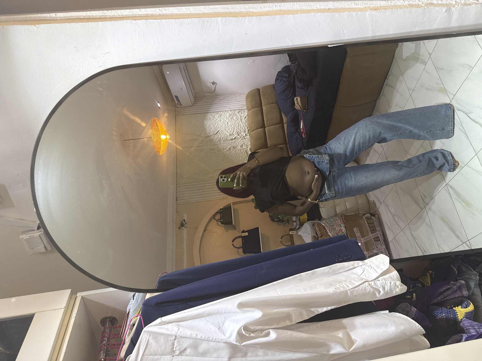 The image shows a person standing in front of a full-length arched mirror, taking a selfie with a phone. The person is wearing a black crop top and light blue flared jeans. Their belly is exposed. In the mirror's reflection, we see a room with a bed covered in a dark blue comforter, a headboard made of brown tufted upholstery, and an air conditioning unit mounted on the wall. There's also a decorative light fixture with a woven orange shade hanging from the ceiling, and a textured wall. To the left of the mirror, a portion of a closet is visible, with several garments hanging on hangers, including a white shirt and a dark blue jacket. The floor is tiled with a light-colored marble pattern. The overall scene suggests a bedroom or dressing area. The image appears to be taken in Abuja, Nigeria, based on the provided context.