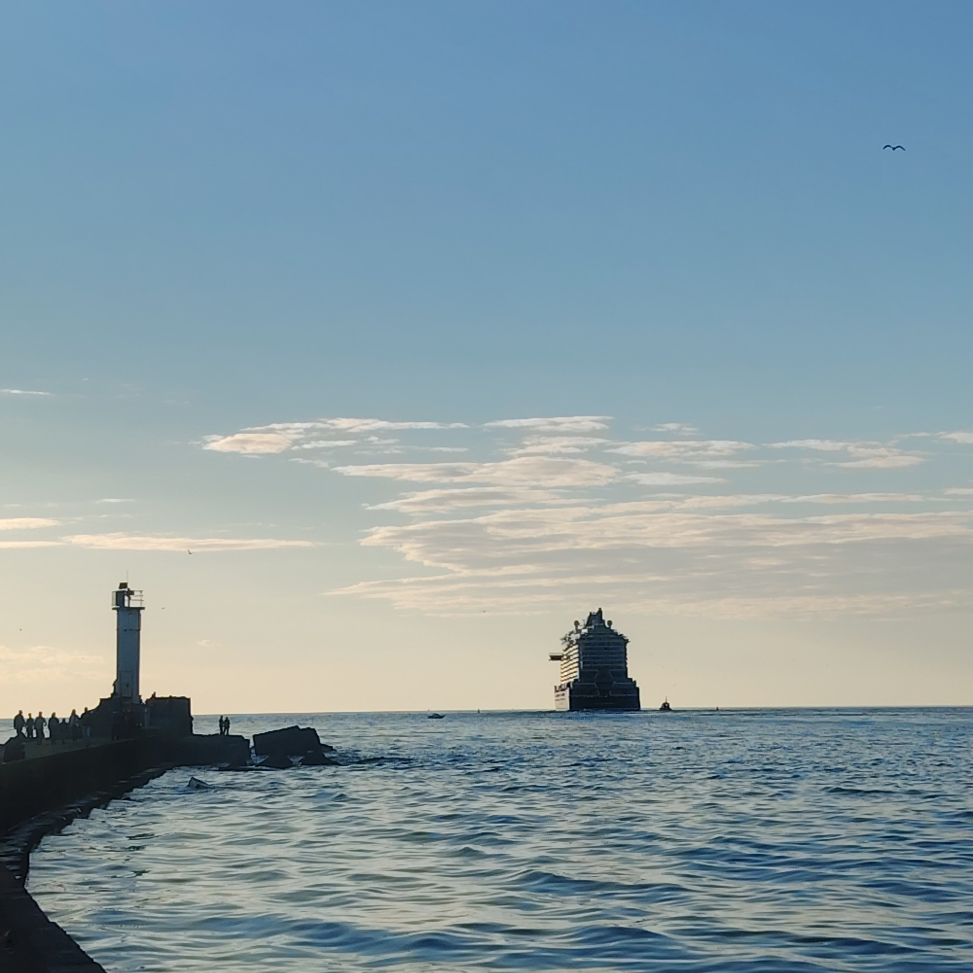 The image depicts a seascape at Riga, Latvia, featuring a lighthouse on a pier in the foreground on the left, a large cruise ship at sea in the background, and a bright blue sky with light clouds.
The lighthouse, painted white with a dark top, stands at the end of a stone pier. Several people are silhouetted against the sky, standing on the pier near the lighthouse.
To the right of the pier, in the sea, sits the large cruise ship at a distance. The ship is dark in color. A couple of smaller boats are around the ship, acting as tenders.
The sea has gentle waves and a gradient of blue and grey colors.
The sky is bright blue with a scattering of thin white clouds. A bird is visible flying in the sky.