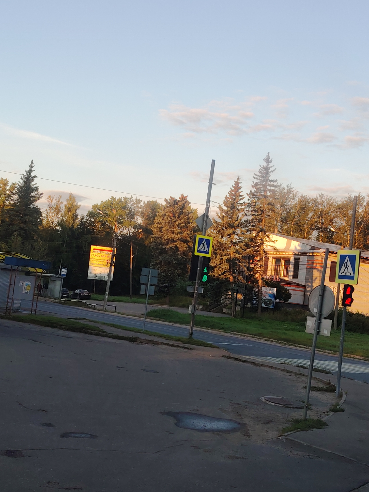 The image depicts a street scene in Konakovo, Russia. In the foreground, a paved road shows several puddles. To the right, a pedestrian crossing is marked, with traffic lights and pedestrian crossing signs. One traffic light displays a red signal, while another shows a green signal. A bus stop shelter is visible to the left, with two individuals standing nearby. Further back, a building with white and orange trim and several trees line the street. The sky above is light blue with scattered clouds, and sunlight casts long shadows, indicating it is likely late afternoon or early evening.
