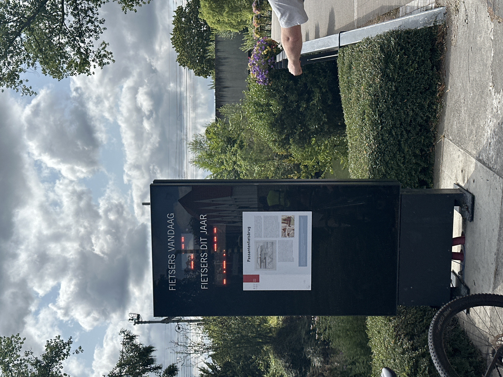 The image depicts a digital display board situated outdoors, likely in a public area. The board displays text in Dutch, with "FIETSERS VANDAAG" (Cyclists Today) and "FIETSERS DIT JAAR" (Cyclists This Year) prominently featured. Below this, there is a poster with an illustration and text titled "Passantenfietsbrug" (Pedestrian-cycle bridge). The surrounding environment is green and lush, with trees, bushes, and a body of water visible. The sky is partly cloudy. A bicycle is partially visible in the foreground, and a person's arm is extending from the right side of the frame, touching a railing. The scene suggests an informational display related to cycling in Bruges, Belgium, possibly near a bridge.