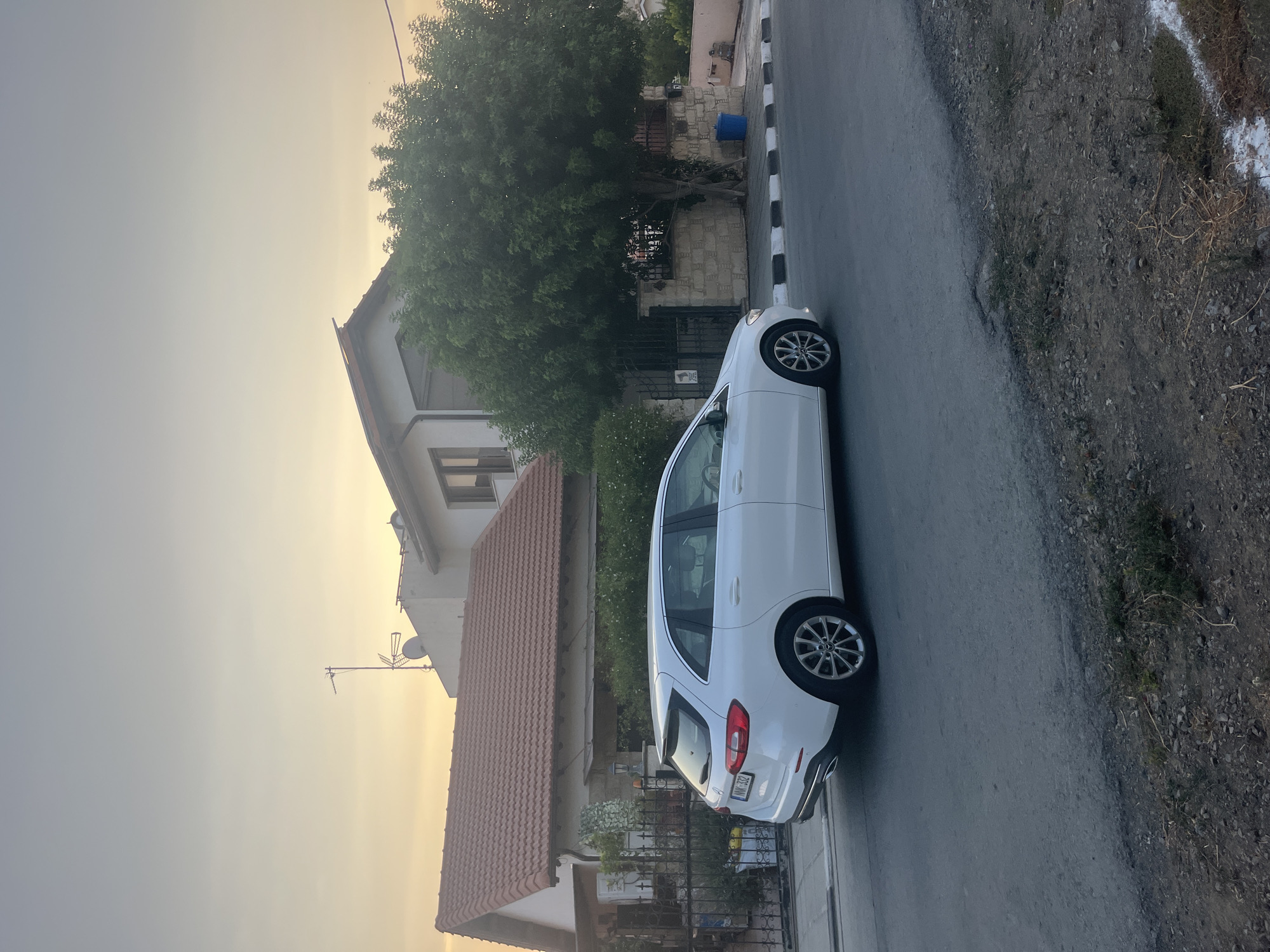 A white Mercedes-Benz B-Class wagon is parked on the side of a paved road in the Parekklisia Community, Cyprus. The car faces right, its rear bumper and rear wheel clearly visible. The license plate is visible and reads "NNK-332". The scene is set during twilight, with a soft, yellowish glow in the sky. A house with a red-tiled roof is visible behind the car, partially obscured by a large, leafy green tree. A black metal fence runs along the front of the property. A blue trash bin is visible on the sidewalk next to the tree. The road surface appears to be asphalt, with a gravelly shoulder. The overall atmosphere is peaceful and suburban.
