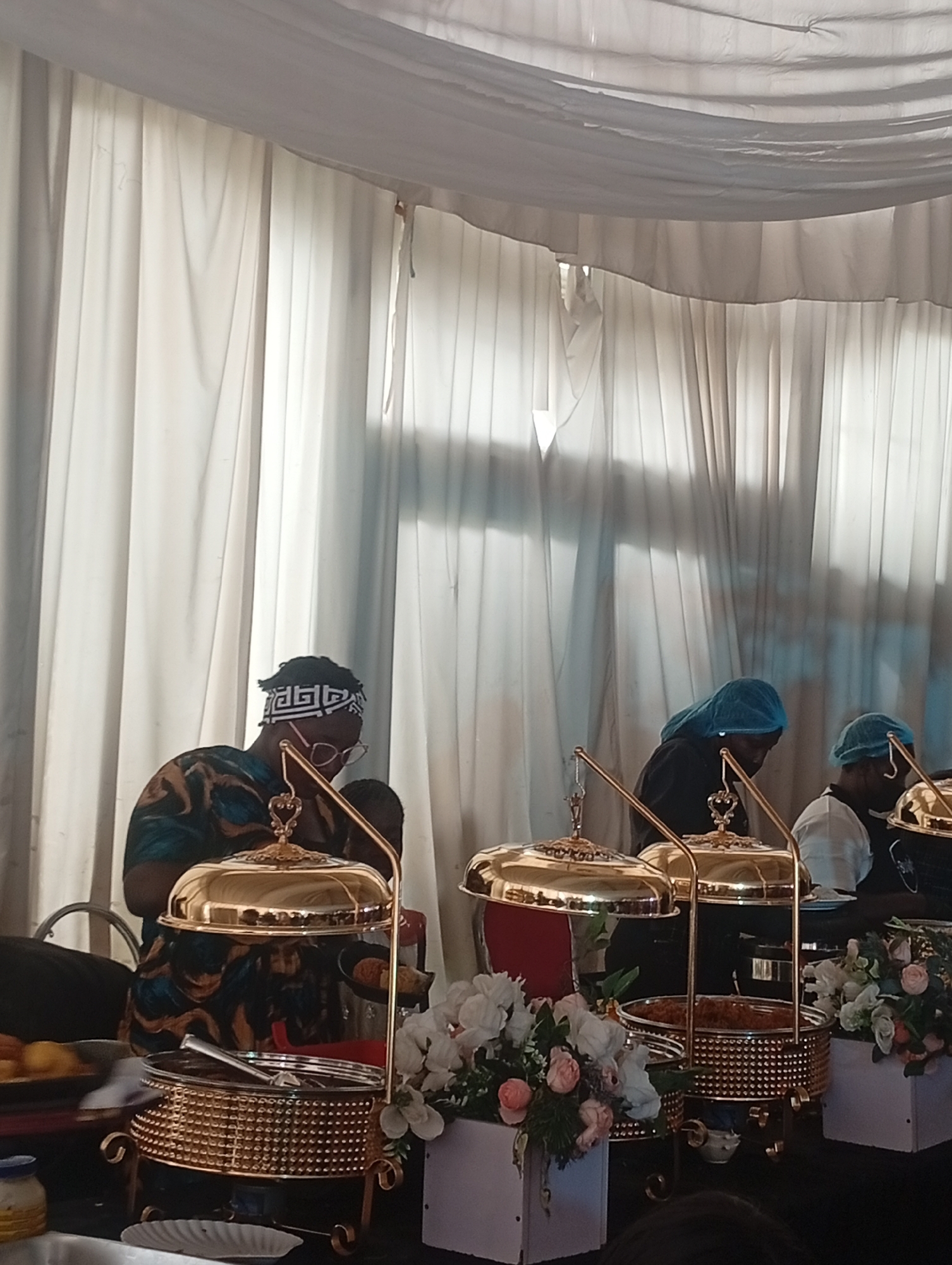 The image shows a catered event, possibly a wedding, in what appears to be an indoor venue in Barakin Lerawi, Nigeria. White drapery serves as a backdrop. Several individuals are seen tending to gold-colored chafing dishes arranged on a black table. One individual wears a patterned shirt and a headband, along with eyeglasses. Two other individuals are wearing blue hairnets. Bouquets of white and pink flowers are interspersed among the chafing dishes. Some plates are on the table. Food service utensils can be seen. The lighting is a mix of natural and artificial.