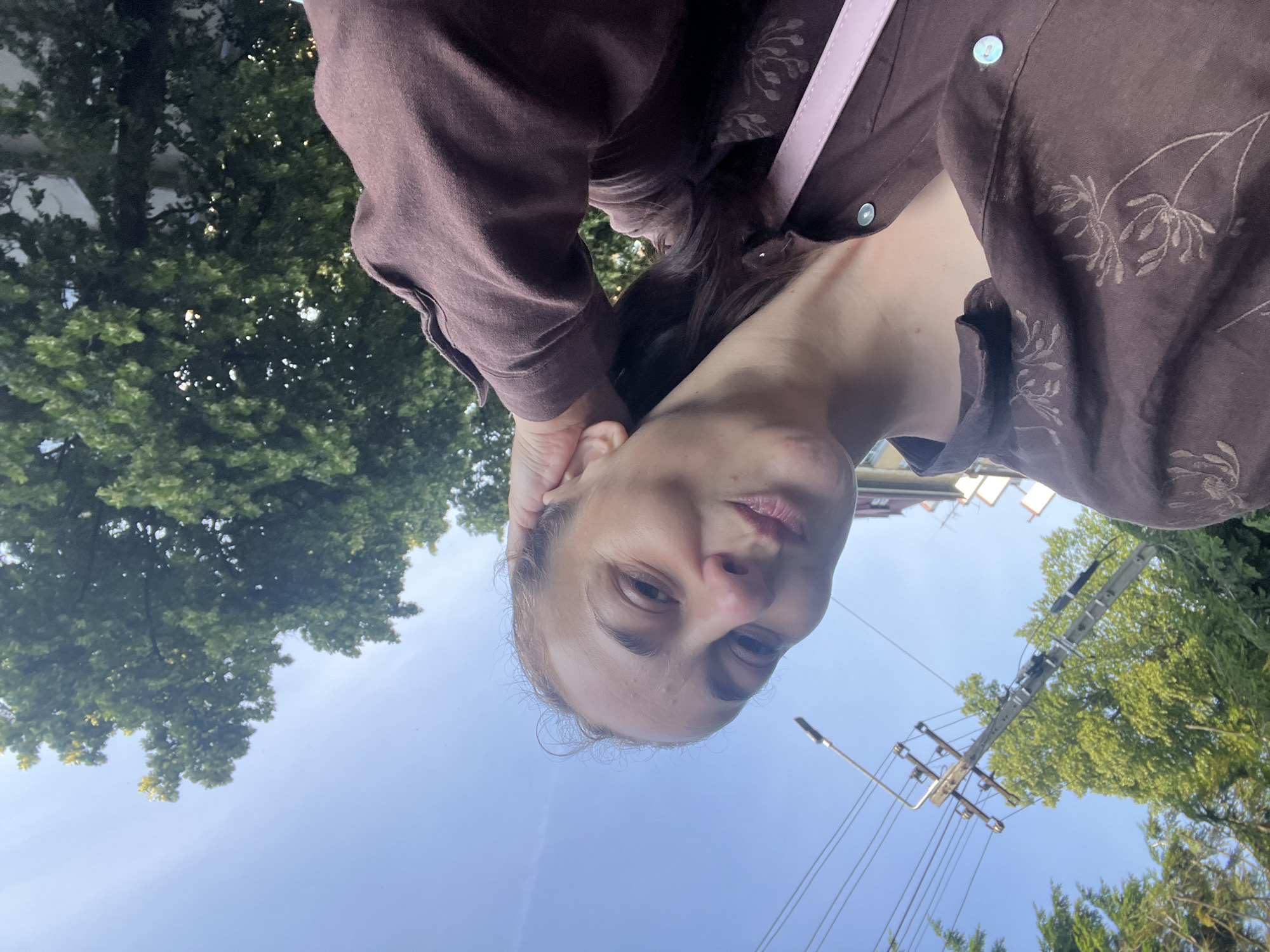 A woman is taking a selfie outdoors on a clear day. She is wearing a brown shirt with a floral pattern and has her right hand resting on her head. Behind her, there are trees and a utility pole with wires. The woman is looking at the camera with a slight smile.