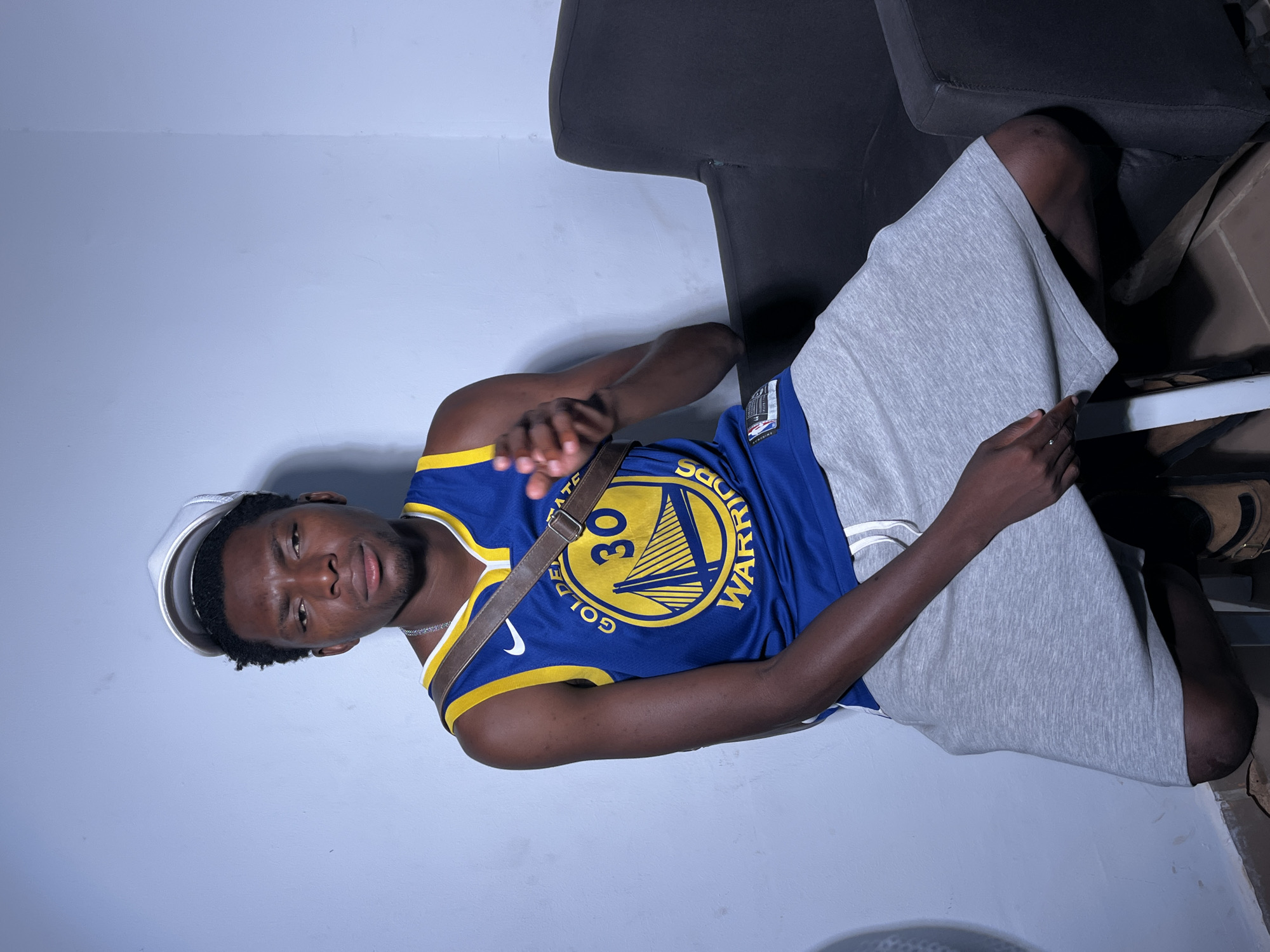 A young Black man wearing a white baseball cap, a blue and gold Golden State Warriors jersey with the number 30, and grey shorts sits on a white stool next to a black armchair. He is leaning to his right and looking towards the camera with a serious expression. His right arm is bent at the elbow and he is gesturing with his right hand, which is partially visible. A brown strap from a bag crosses his chest over the jersey. The background is a plain light blue wall. The lighting is harsh and creates shadows. The scene appears to be indoors, possibly in an apartment or studio.