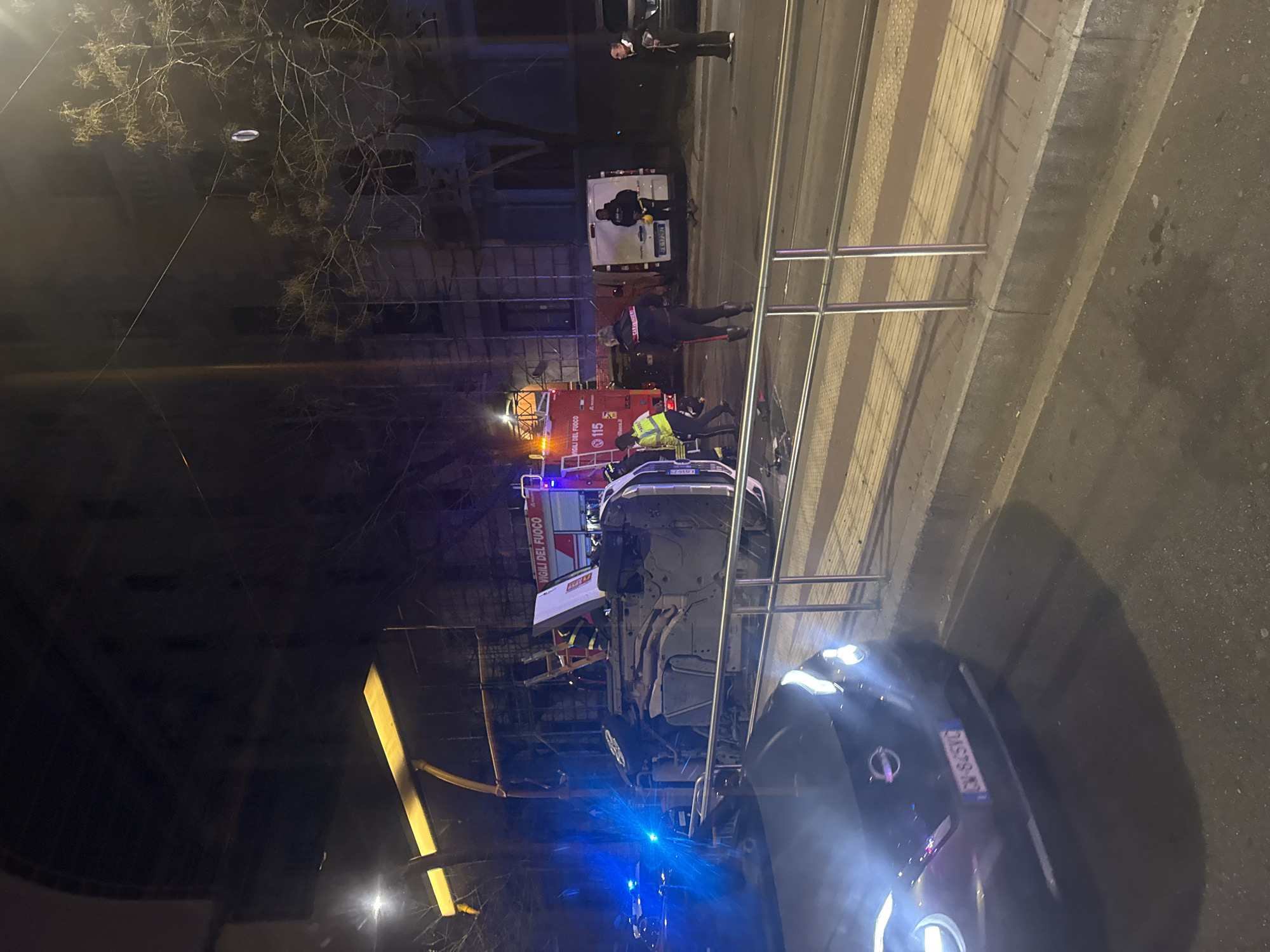 The image depicts an urban nighttime scene in Milan, Italy, showing the aftermath of a vehicular incident involving an overturned white car.
In the center of the frame, a white vehicle is lying on its roof, resting on a raised median or tram tracks. The underside of the vehicle is fully exposed. Adjacent to the overturned car, a red "VIGILI DEL FUOCO" (Italian Fire Department) truck, marked with "115," is parked with active flashing red and blue lights. Several emergency personnel are present; at least two firefighters in uniform, one wearing a high-visibility yellow jacket, are near the overturned vehicle. A Carabinieri officer, identified by "CARABINIERI" on their back, stands near the incident. Another emergency vehicle with flashing blue lights is partially visible in the left foreground.
Further down the street to the right, a white van with license plate "FE 172EN" is parked. Another uniformed individual in a dark outfit with a yellow high-visibility vest is near this van. A male civilian in a dark jacket is observing the scene from the sidewalk.
In the immediate foreground, a dark-colored vehicle, identifiable as a Nissan by its emblem, has its headlights illuminated, facing away from the camera. Its Italian license plate reads "CW 845VC."
The environment includes multi-story buildings in the background, one of which is partially covered in scaffolding. Bare trees line the street. Overhead wires are visible. The foreground features an asphalt road separated by a metal-railed barrier from a raised sidewalk or tram platform with tactile paving. Street lighting and vehicle lights provide illumination for the nighttime setting.